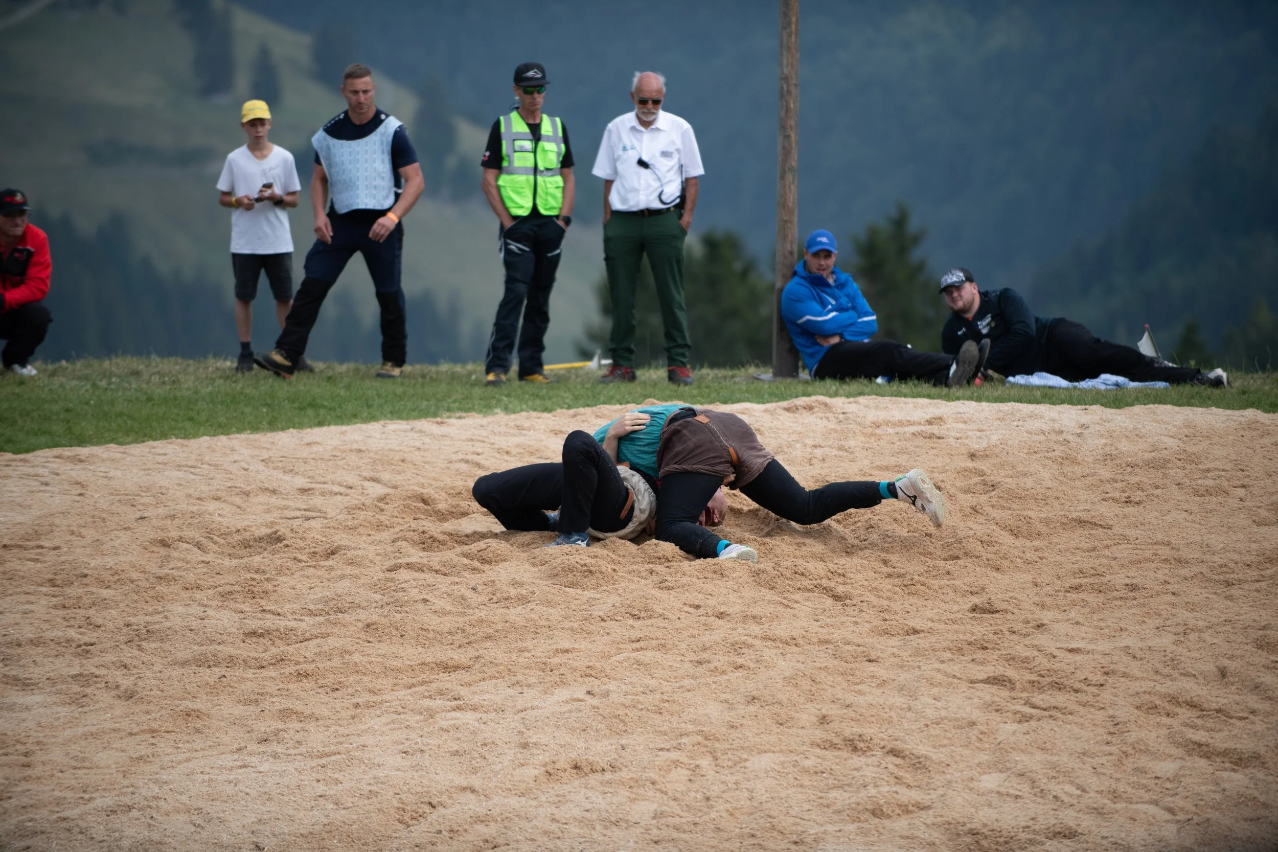 Menschen beobachten einen Unfall auf einer Sandbahn, bei dem eine Person auf dem Boden liegt und eine weitere Person with Hands on Kopf nach dem Sturz auf dem Boden liegt, während andere Personen zuschauen.