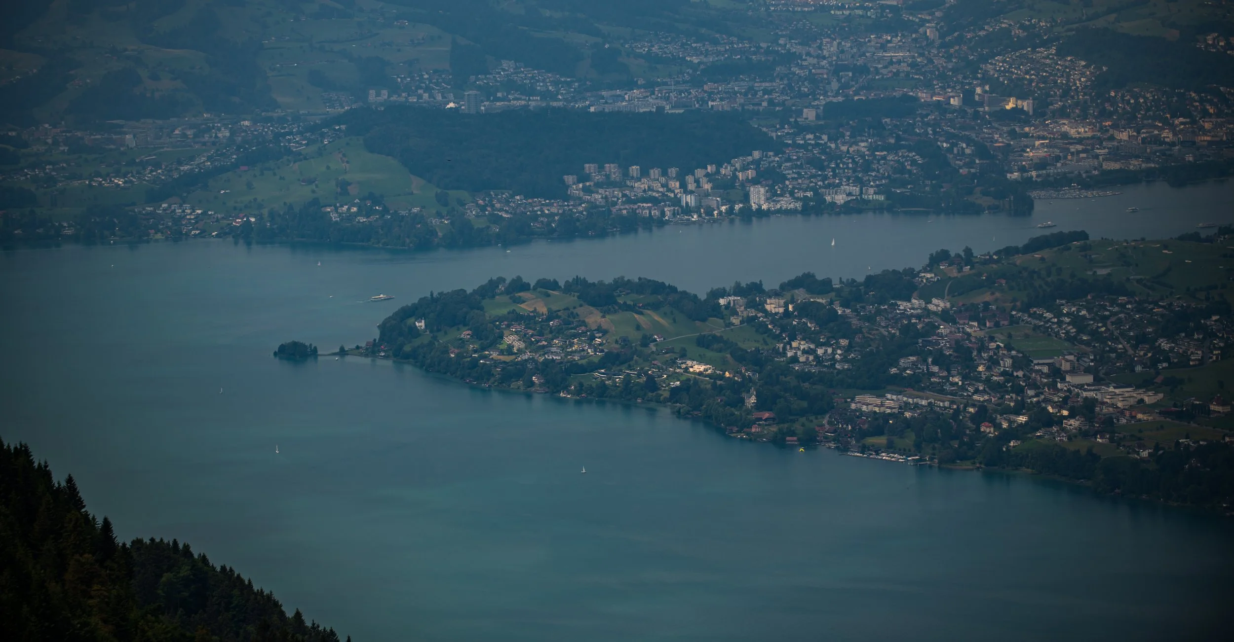 Luftaufnahme eines Sees mit umgebenden Stadtvierteln, Hügeln und bewaldeten Bereichen, bei leicht bewölktem Himmel.