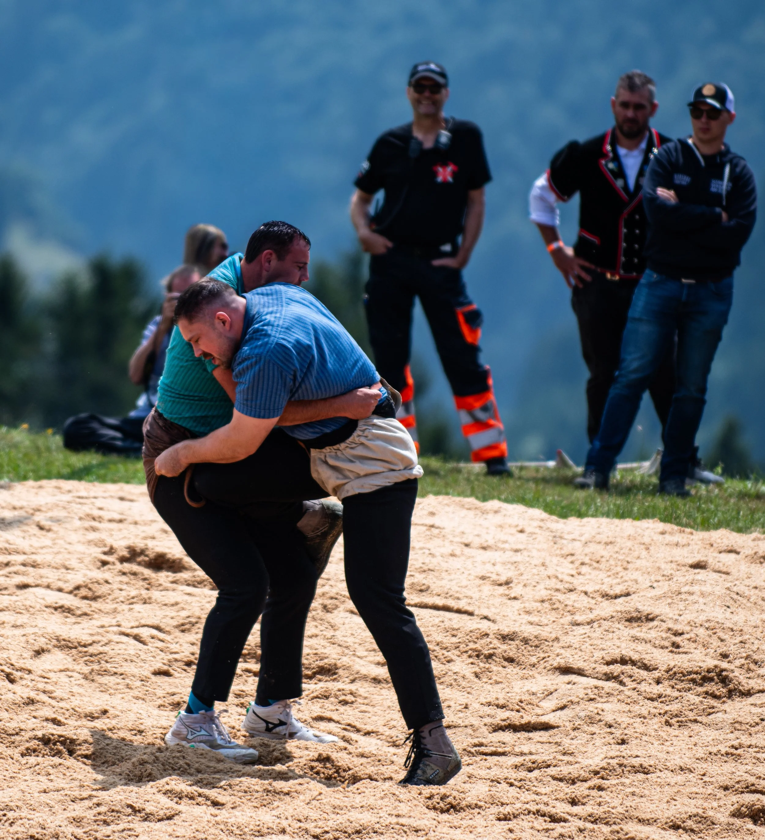 Zwei Männer in schwarzer Kleidung kämpfen auf einem Sandplatz, während andere Männer im Hintergrund zuschauen, bei solchen Aktivitäten.