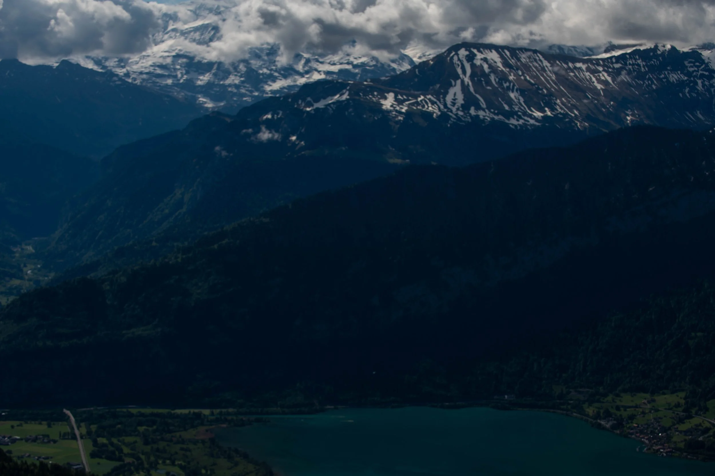 Berglandschaft mit schneebedeckten Gipfeln, dunklen Hängen und einer Seenlandschaft im Vordergrund, bewölkter Himmel.