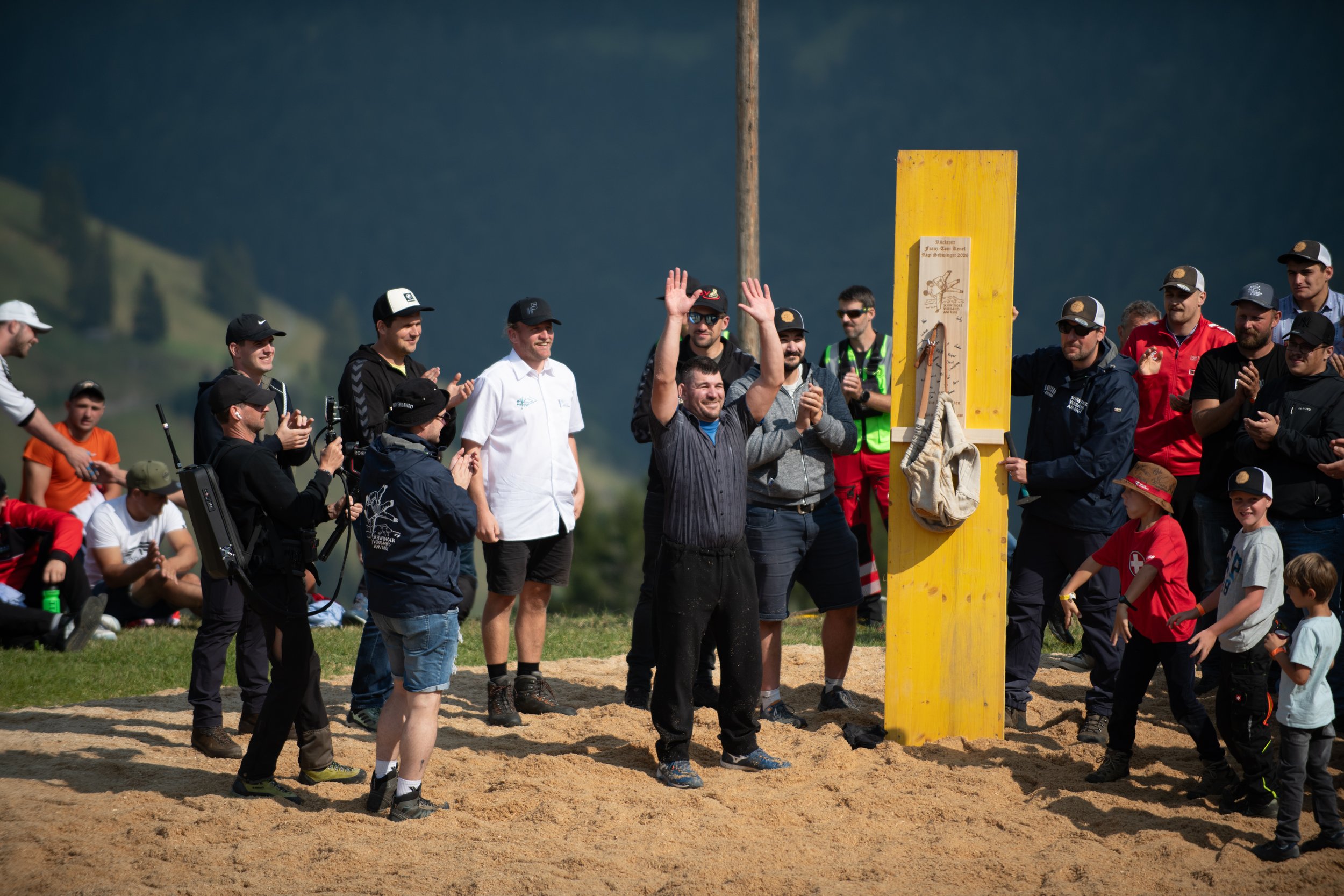 Menschen versammeln sich bei einem Wettbewerb im Freien, einige klatschen und feiern. Im Mittelpunkt steht ein Mann, der gerade einen Pokal hält, während andere zuschauen und applaudieren.