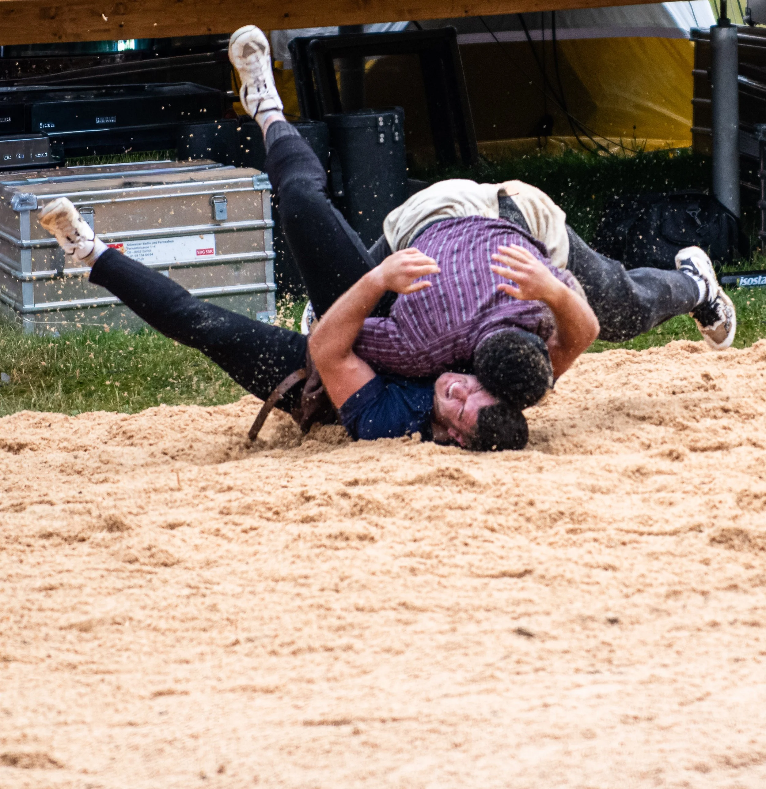 Zwei Männer liegen auf dem Boden, einer hält den anderen fest, während im Hintergrund jemand mit ausgestrecktem Bein auf den Boden tritt, alles auf einer Sandfläche.