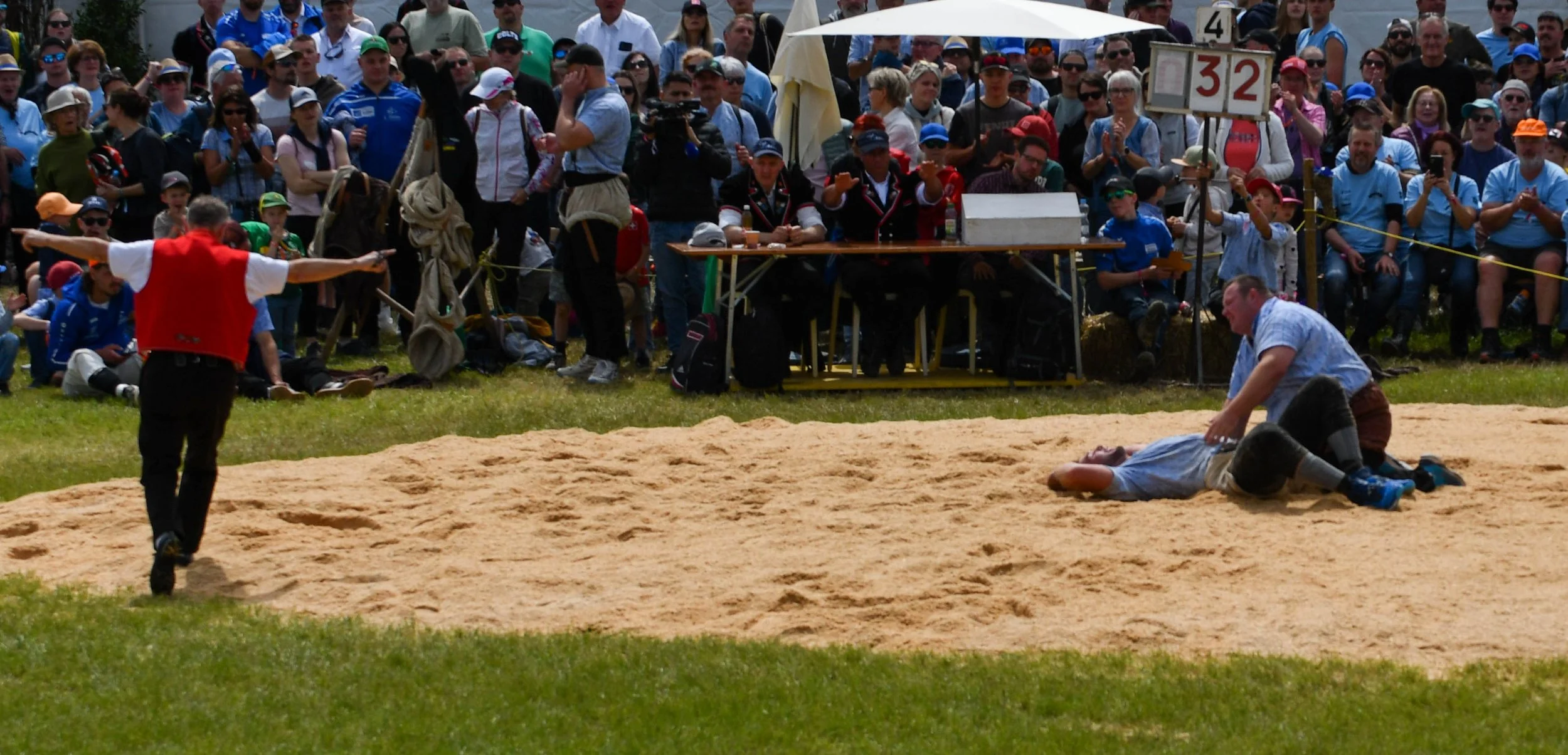 Wrestling match auf Sand, mit einem Betrachterfeld im Hintergrund, inklusive Schiedsrichter und kampfenden Wrestler.