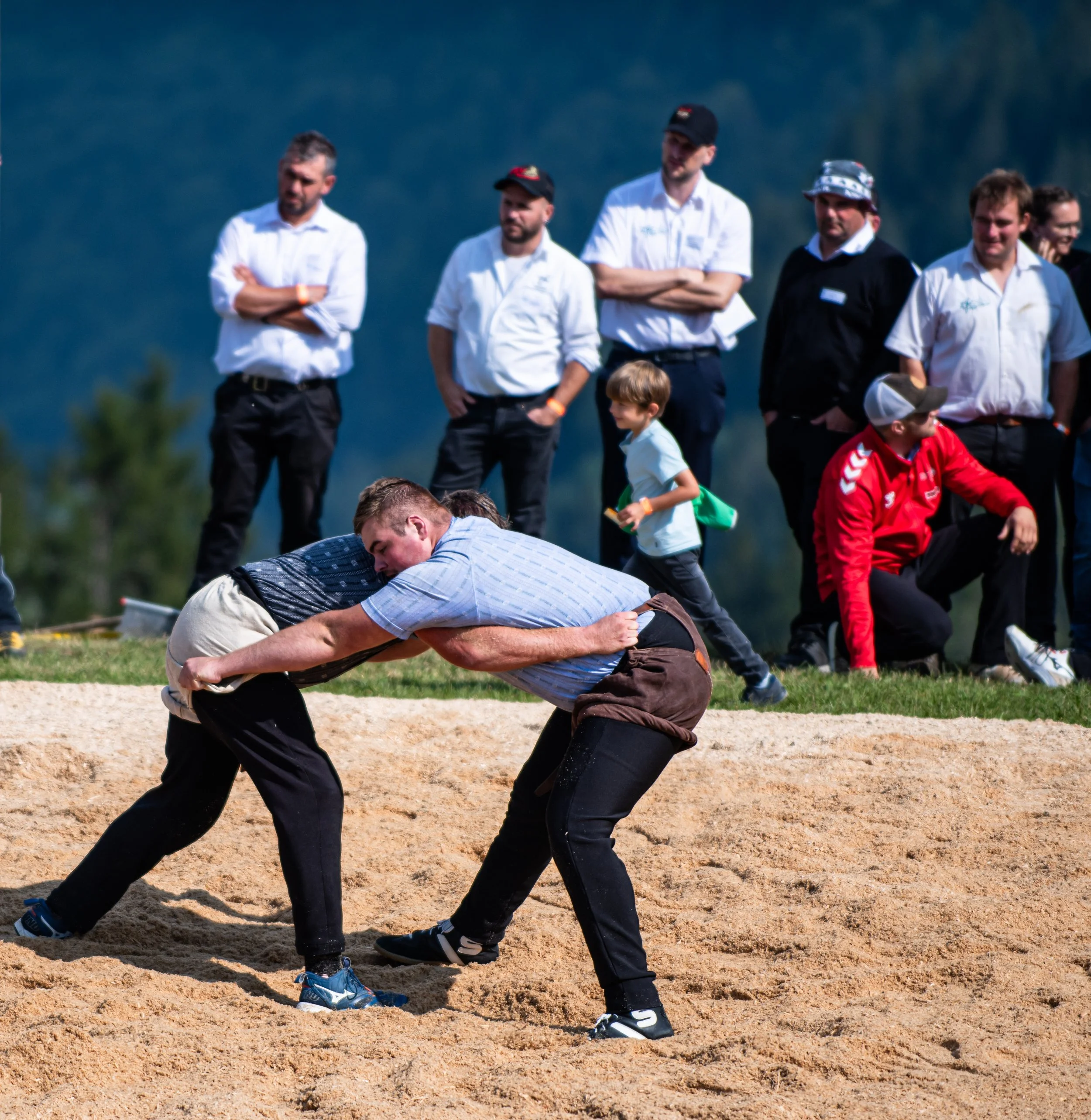 Mehrere Männer in weißen Hemden, einige mit Hüten, stehen auf einer Sandfläche hinter einem Wrestling-Ring. Ein Mann in einem blauen Hemd ist im Vordergrund, er neigt sich nach vorne, während andere zusehen.