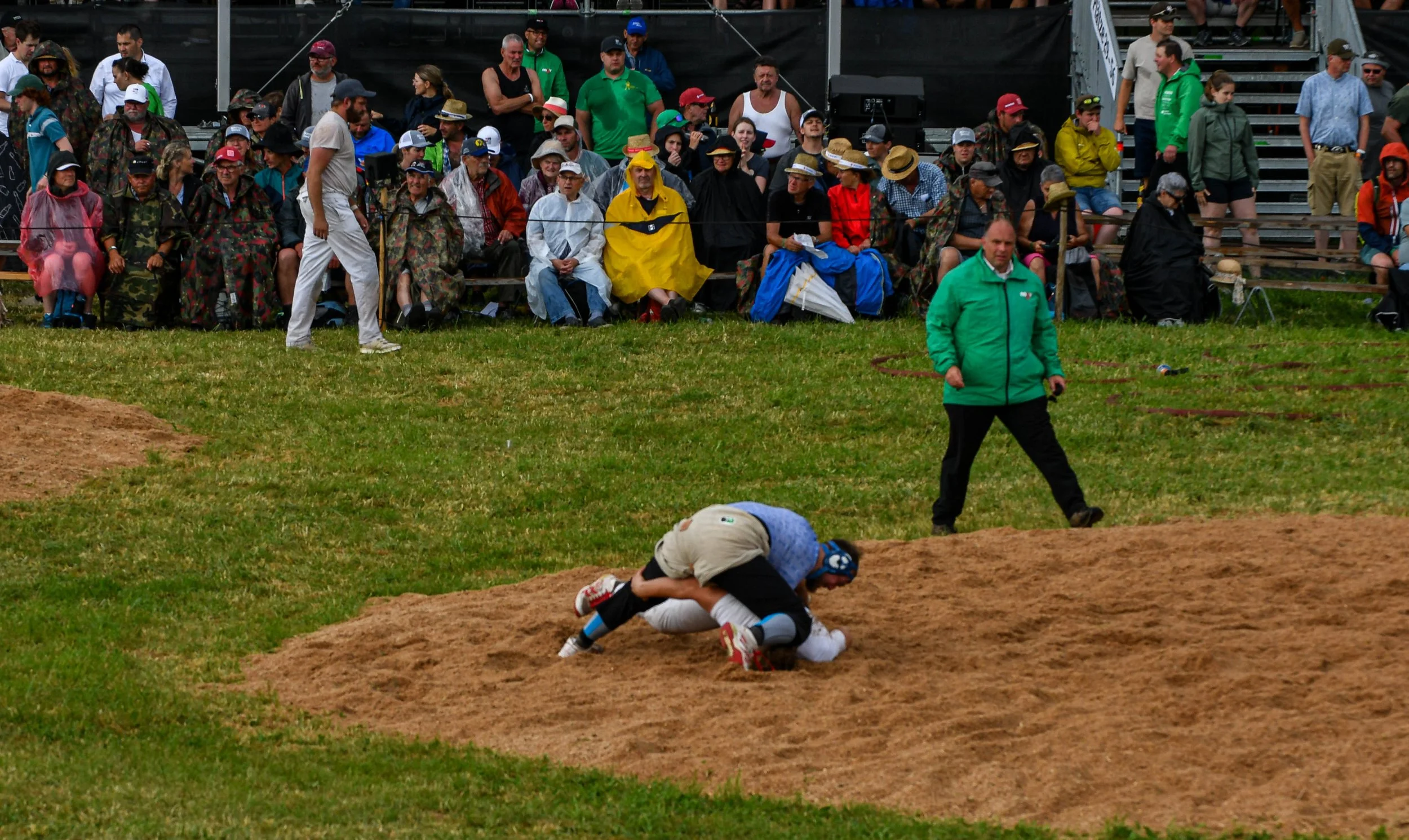Zwei Jugendliche in Sportkleidung beim Ringen auf einem Sandring, im Hintergrund eine Zuschauertribüne mit Menschen, einige tragen Sonnenhüte und Regencapes, während andere im Regen stehen.