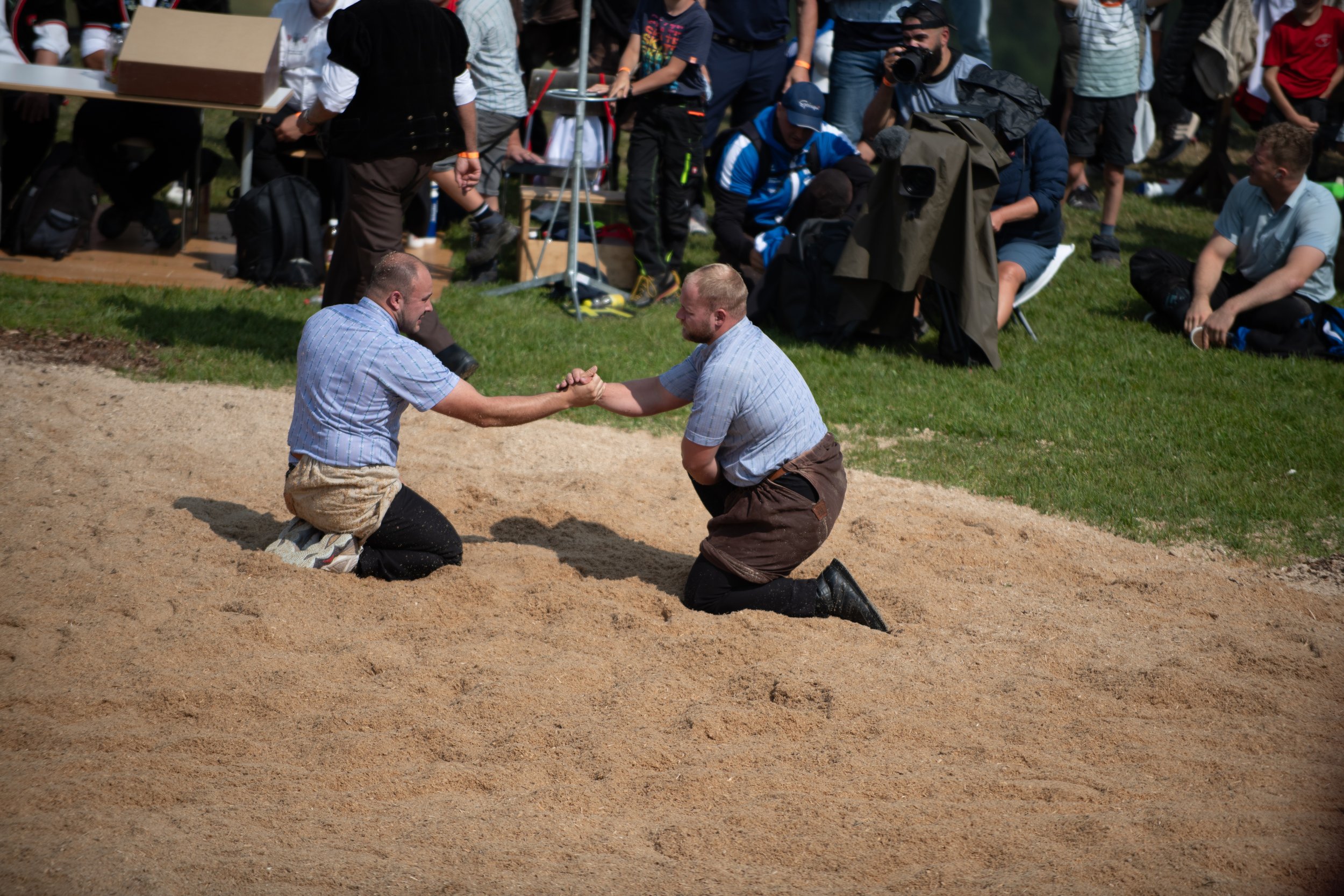 Zwei Männer im Sitzen, die sich die Hände schütteln, während sie in einem Sandkasten knieen, im Hintergrund eine Gruppe von Menschen, die zuschauen und fotografieren.