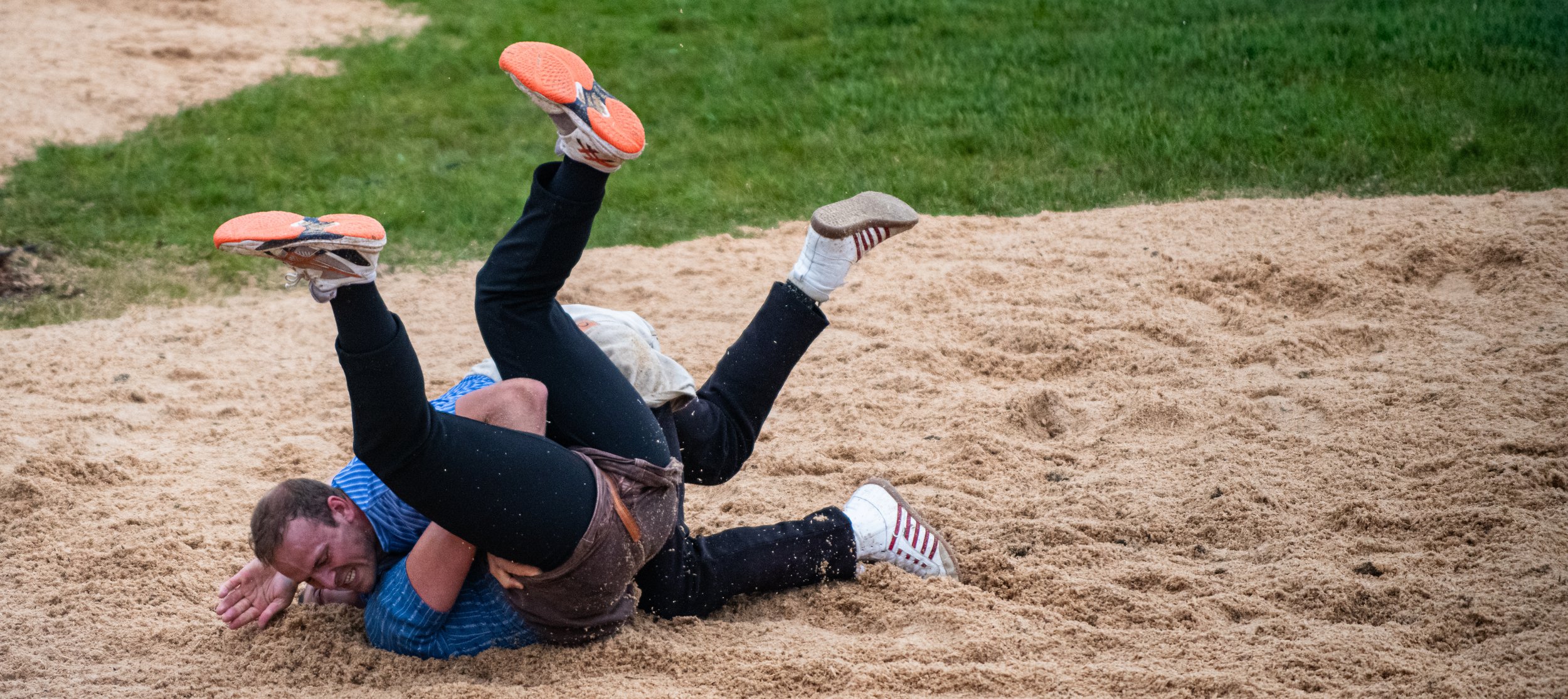 Zwei Männer, die im Sandkasten auf dem Boden liegen und miteinander kämpfen, während sie lachen. Einer trägt eine blaue Jacke und der andere dunkle Kleidung. Im Hintergrund ist Gras zu sehen.