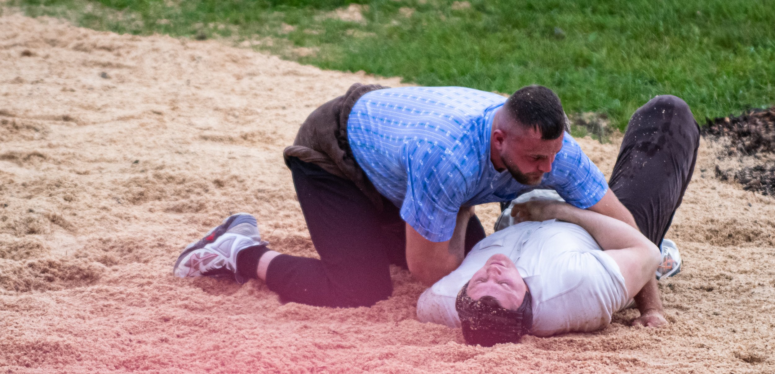 Zwei Männer auf einem Sandplatz bei einem Kampf, einer steht über dem anderen, der auf dem Rücken liegt.
