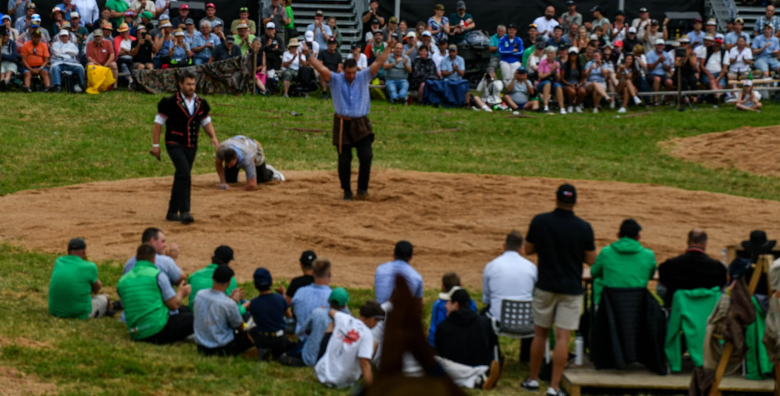 Menschen bei einem traditionellen mexikanischen Rodeo, das auf einer abgesteckten Arena auf Gras stattfindet, mit einem großen Publikum im Hintergrund, das auf Stufen sitzt und zusieht.