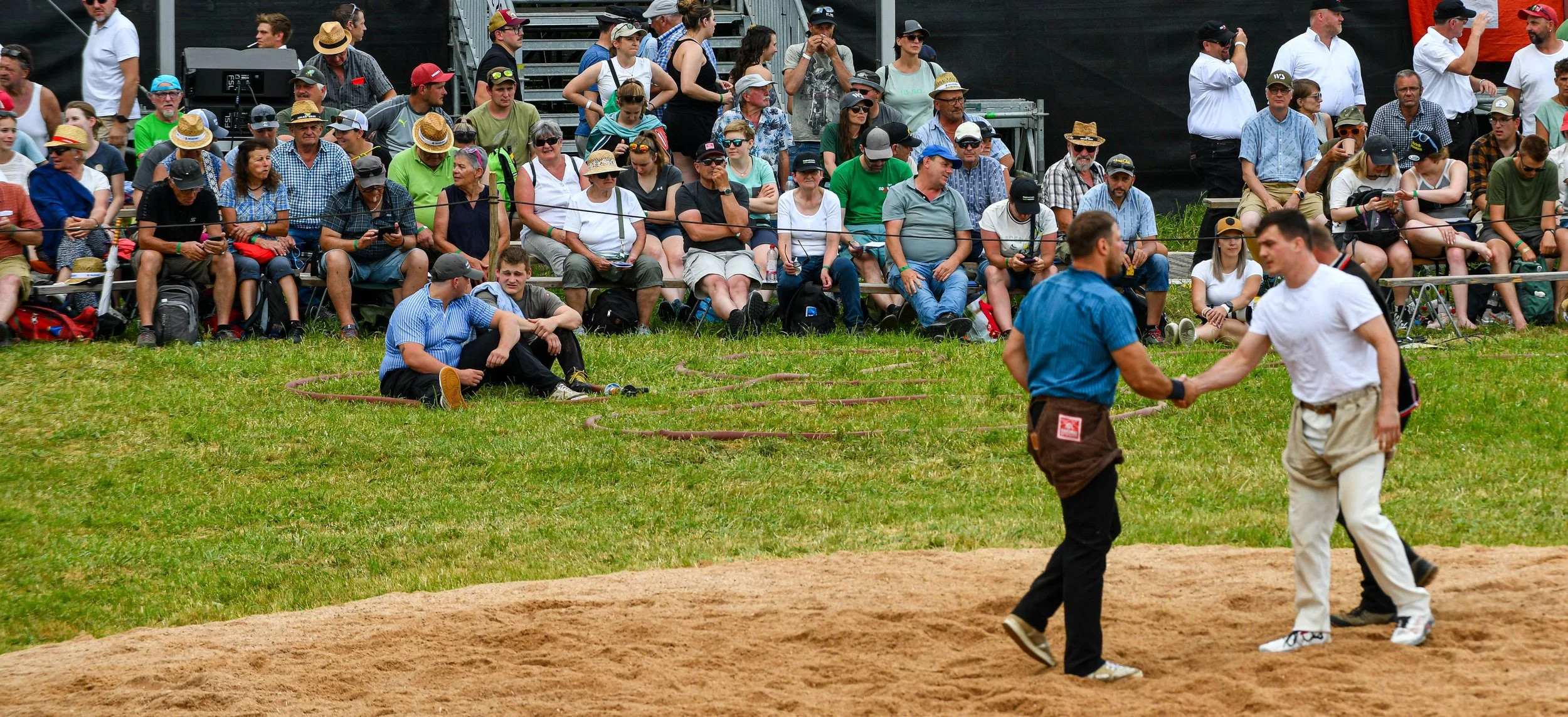 Menschen sitzen auf Bänken und im Gras, einige tragen Hüte und Sonnenbrillen, während zwei Personen im Vordergrund auf einer Sandfläche Händeschütteln, vermutlich bei einem öffentlichen Event oder Festival.