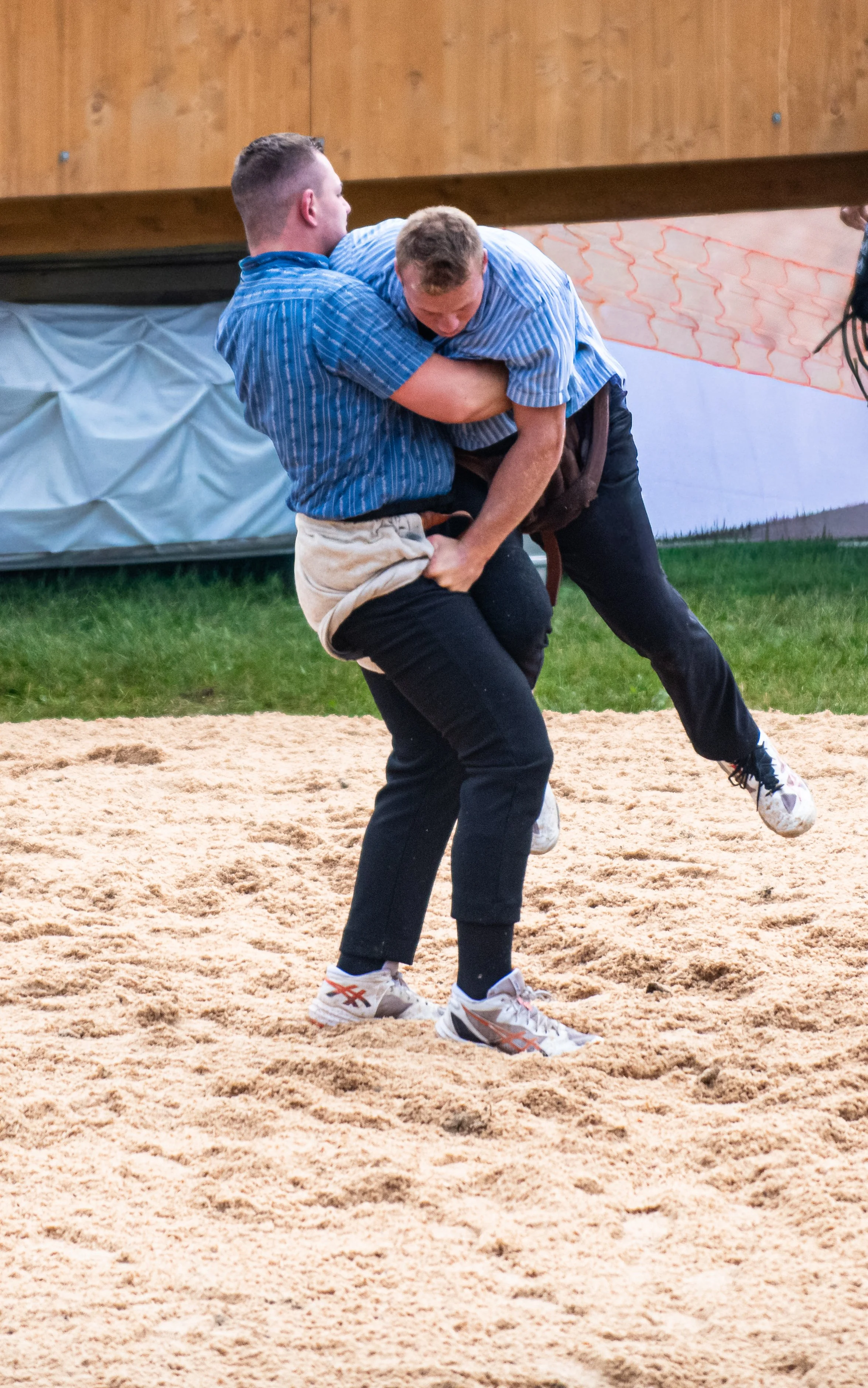 Zwei Männer beim traditionellen bayerischen Schafkopfspiel stehen auf einem Sandboden, einer hebt den anderen hoch, beide tragen blaue Hemden und schwarze Hosen.