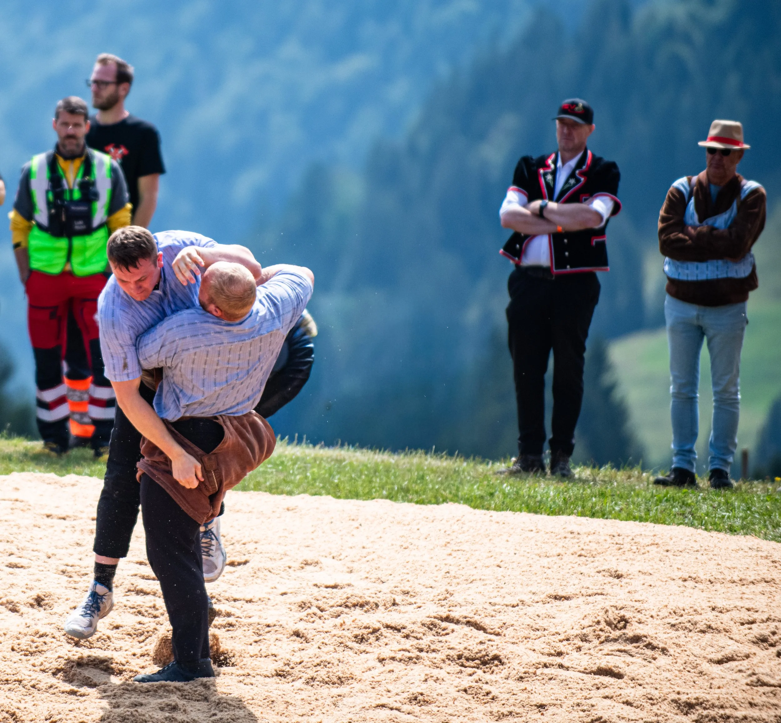 Personen beim Ringen im Sand, während andere Personen im Hintergrund stehen und zuschauen, auf einer grünen Wiese mit einer bewaldeten Bergkulisse im Hintergrund.