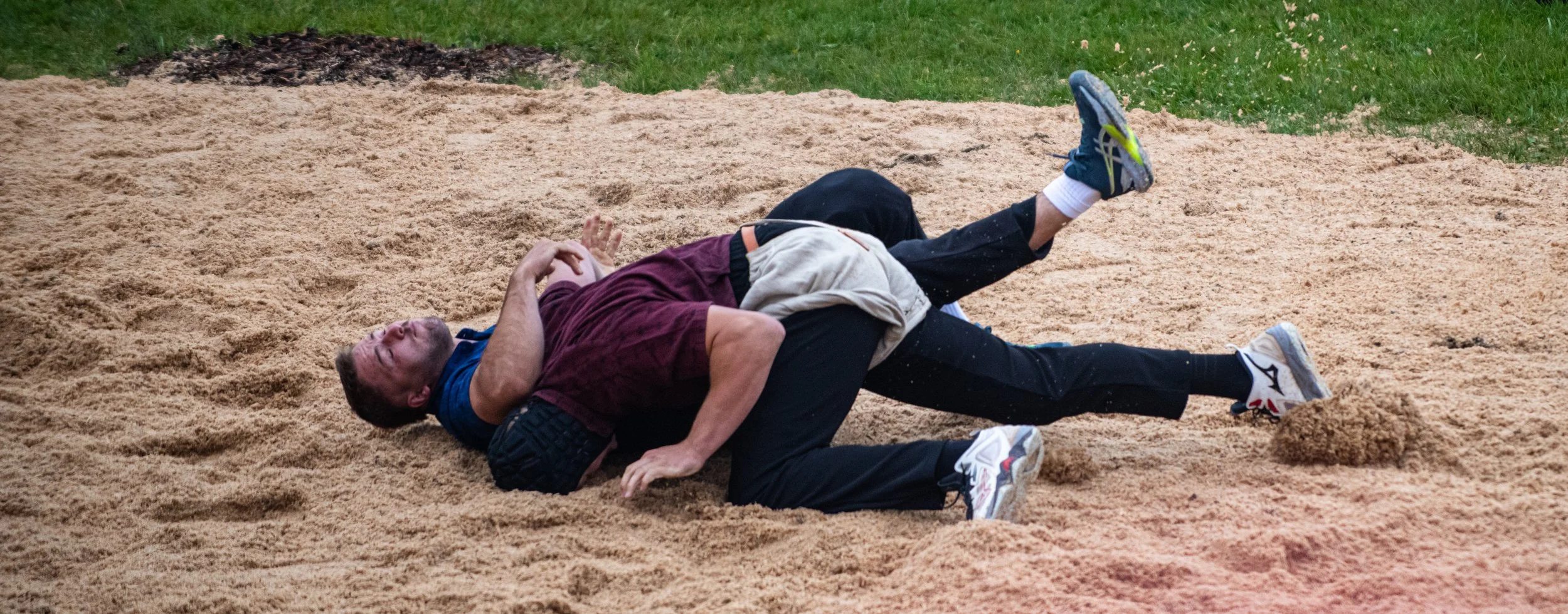 Zwei Männer kämpfen auf einer Sandbahn im Freien. Einer trägt ein blaues T-Shirt und der andere ein maroonfarbenes T-Shirt, beide sind im Grappling/Gegengewichtskampf.