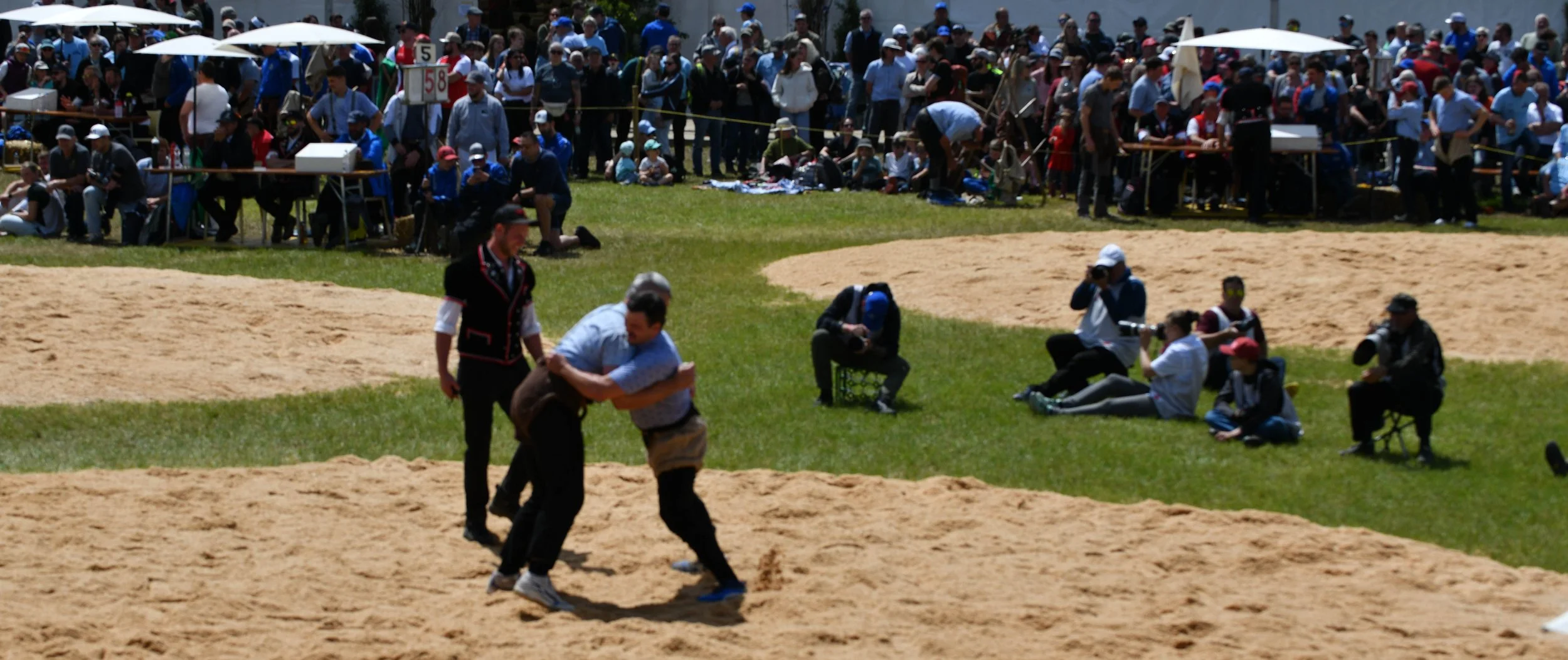 Zwei Männer beim Ringen auf einer Sandfläche während eines öffentlichen Events, mit einem großen Publikum im Hintergrund, das auf Bänken und im Stehen zuschaut.