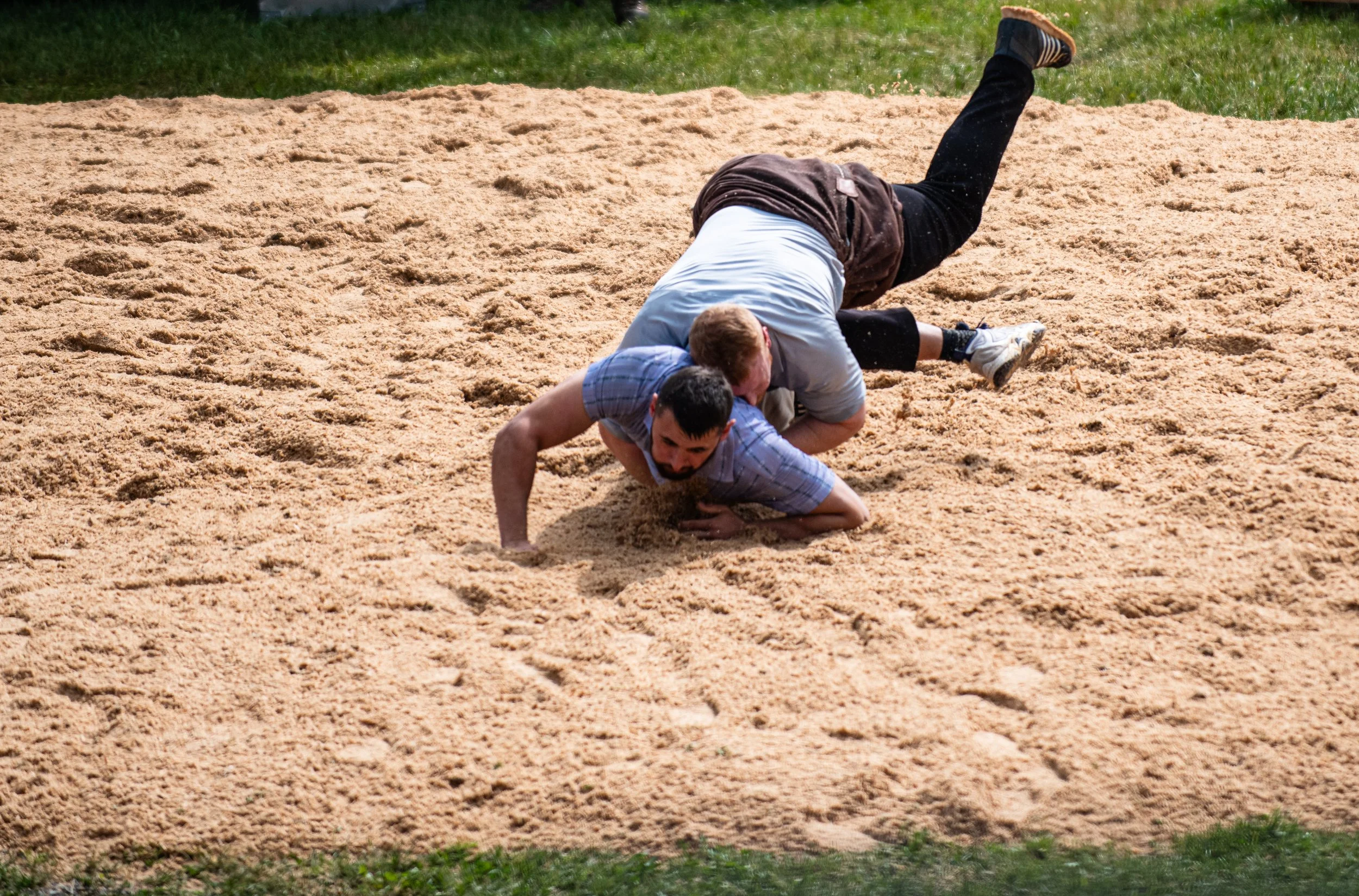 Zwei Männer im Ringen auf einem Sandfeld, einer liegt auf dem Boden, der andere ist oben. Es ist ein Wettkampf oder Kampf im Sand. Im Hintergrund grüne Grasfläche.