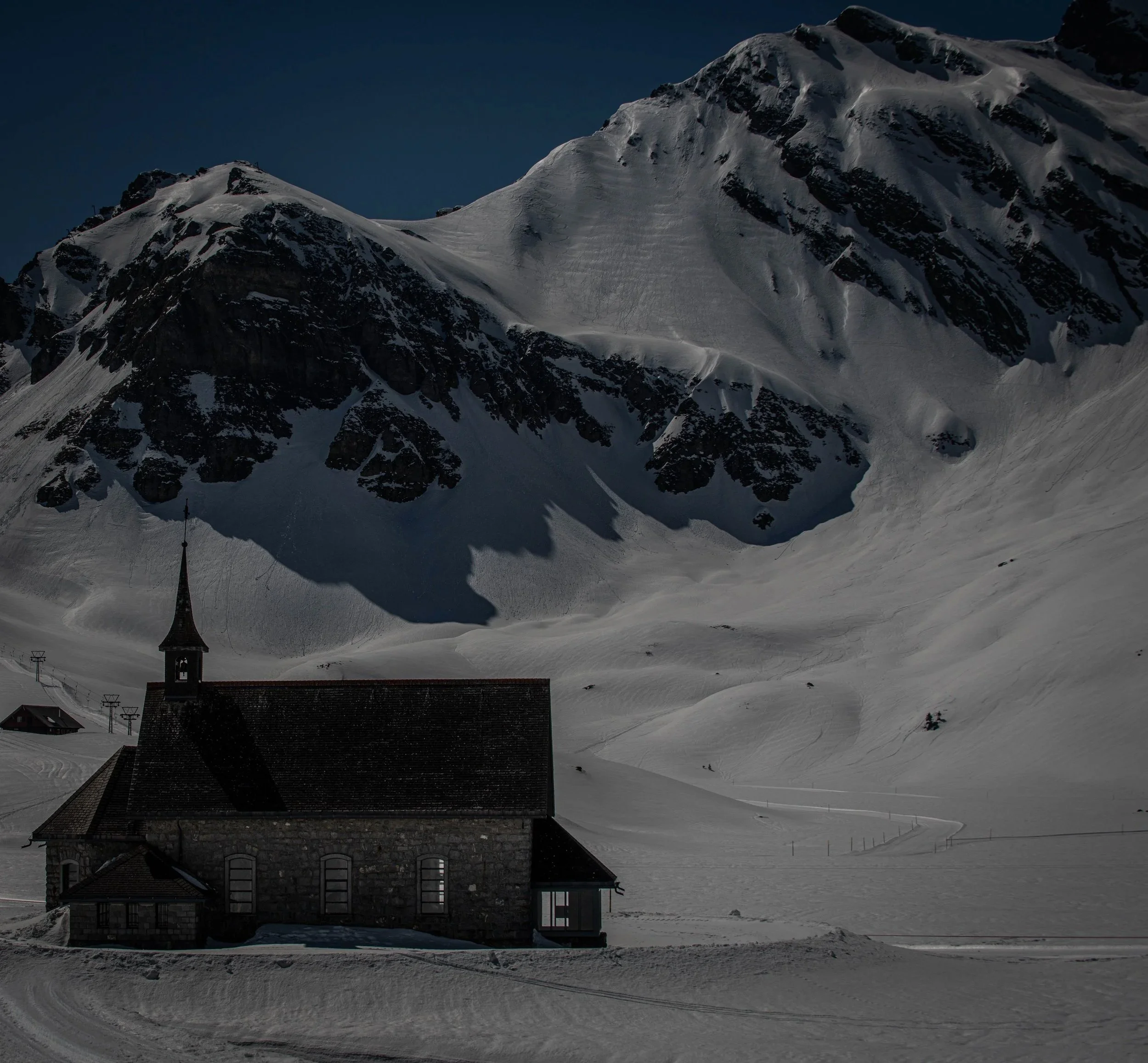Ein schneebedecktes Bergdorf mit dunklem Berggipfel im Hintergrund, darunter ein Haus mit Turm, umgeben von schneebedeckter Landschaft. Klare, dunkle Himmel.