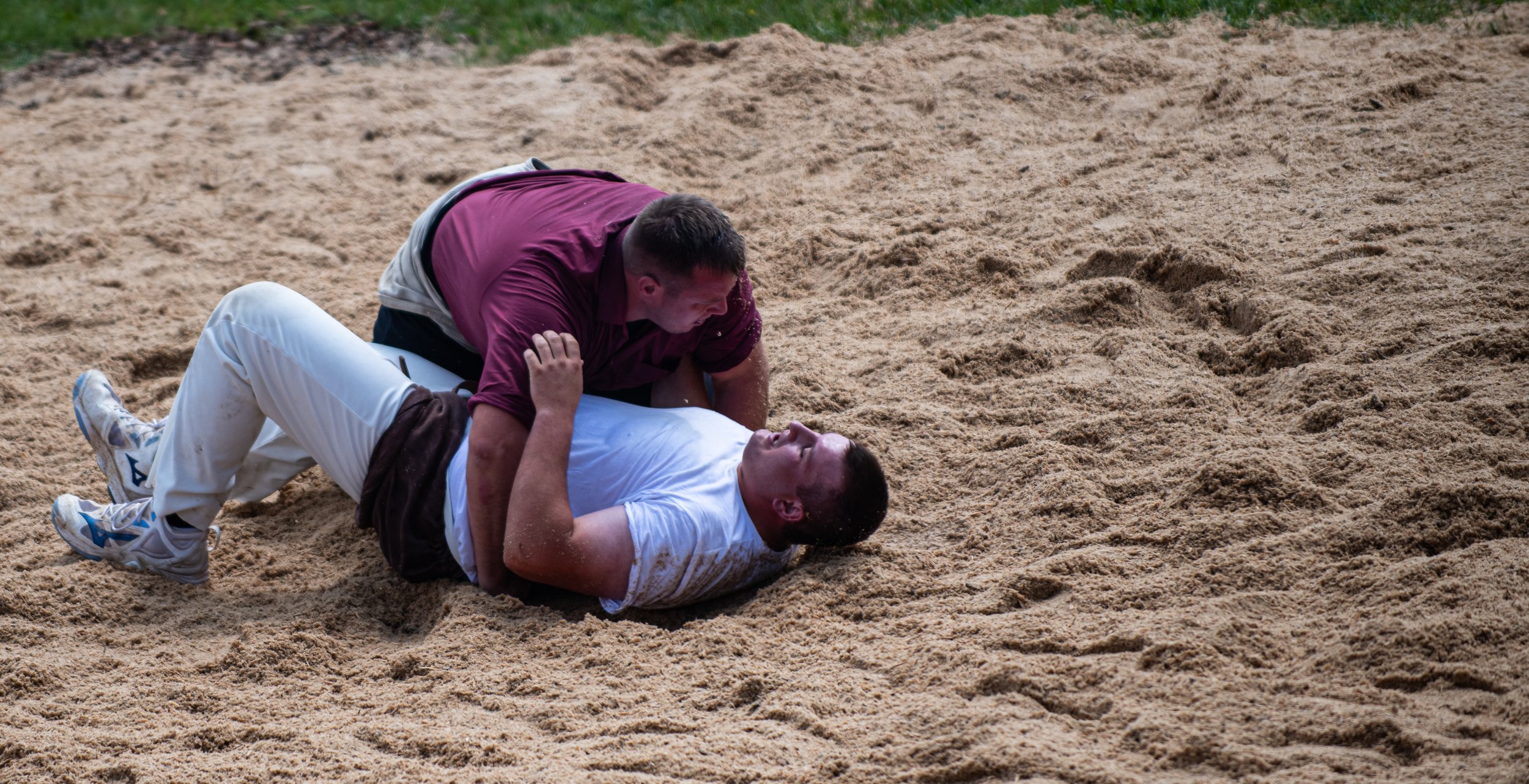 Zwei Männer im Sandkampf, einer liegt auf dem Rücken, der andere sitzt über ihm, beide in Sportkleidung.