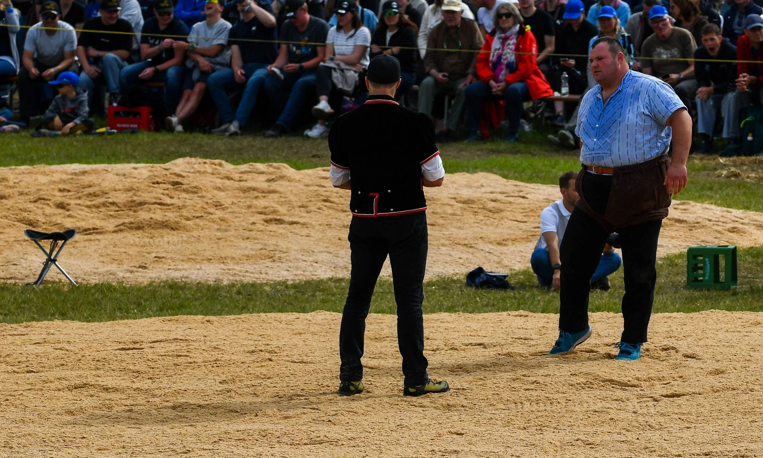 Zwei Männer in traditioneller Kleidung stehen auf einem Sandplatz während eines Events. Im Hintergrund sitzt eine große Menschenmenge, viele tragen Sonnenbrillen und Mützen.