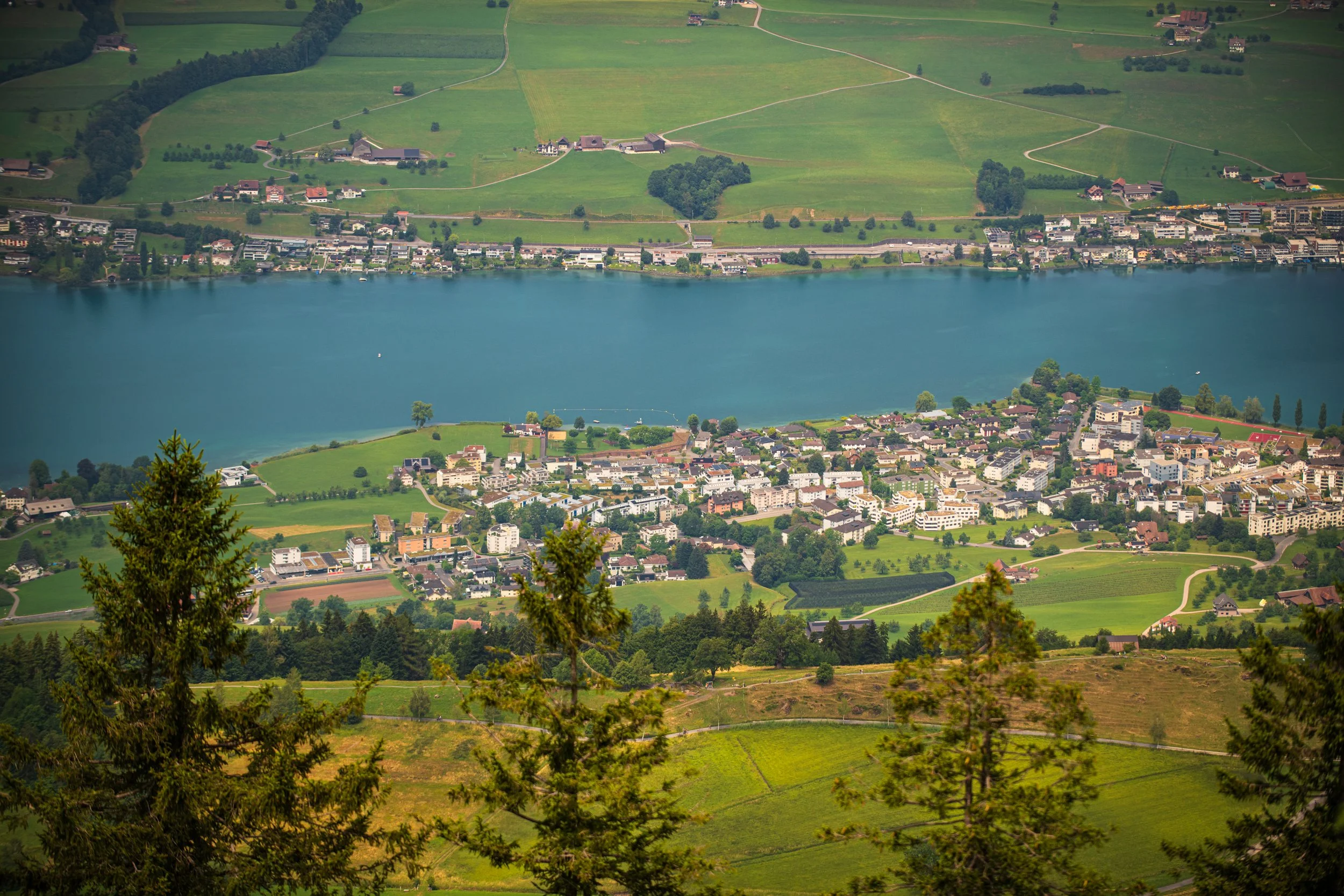 Luftaufnahme eines Sees mit umliegender Wohn- und Landwirtschaftsgebiet, umgeben von grünen Hügellandschaften und Wäldern.