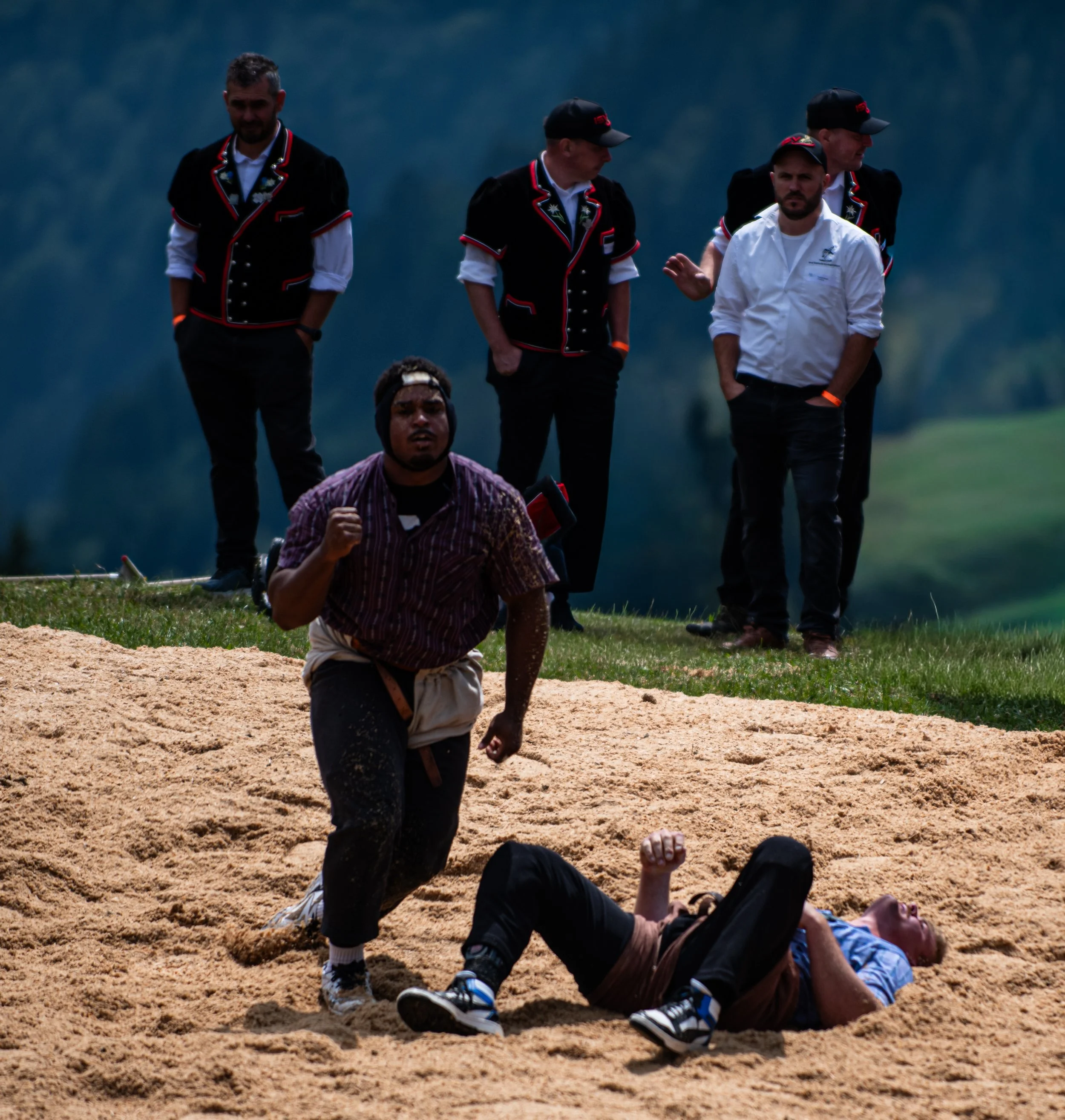 Menschen beim Ringen auf einer Sandfläche im Freien, einige sind in traditionellen schwarzen Kleidern mit roten und weißen Details, eine Person trägt ein weißes Hemd, Hintergründe mit Bäumen und Bergen.