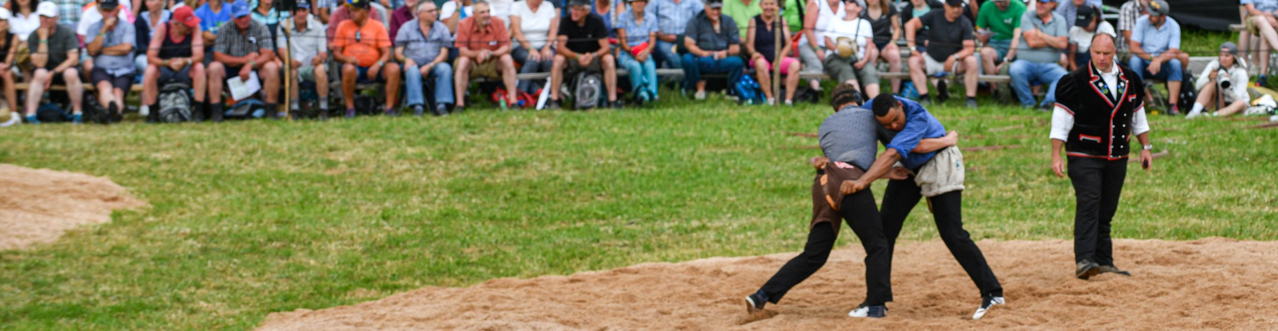 Zwei Männer kämpfen in einem Sandring im traditionellen Ringkampf, während ein Schiedsrichter den Kampf beobachtet. Im Hintergrund sitzen viele Zuschauer.