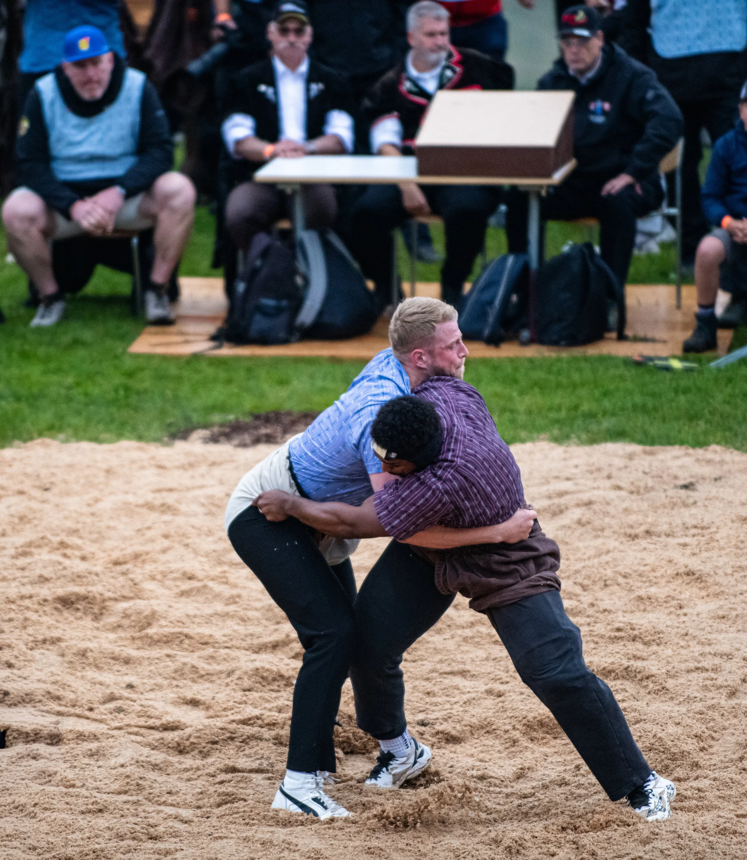 Zwei Männer kämpfen auf einem Sandplatz, die eine greift die andere am Oberkörper fest während eines Wrestlingspiels.