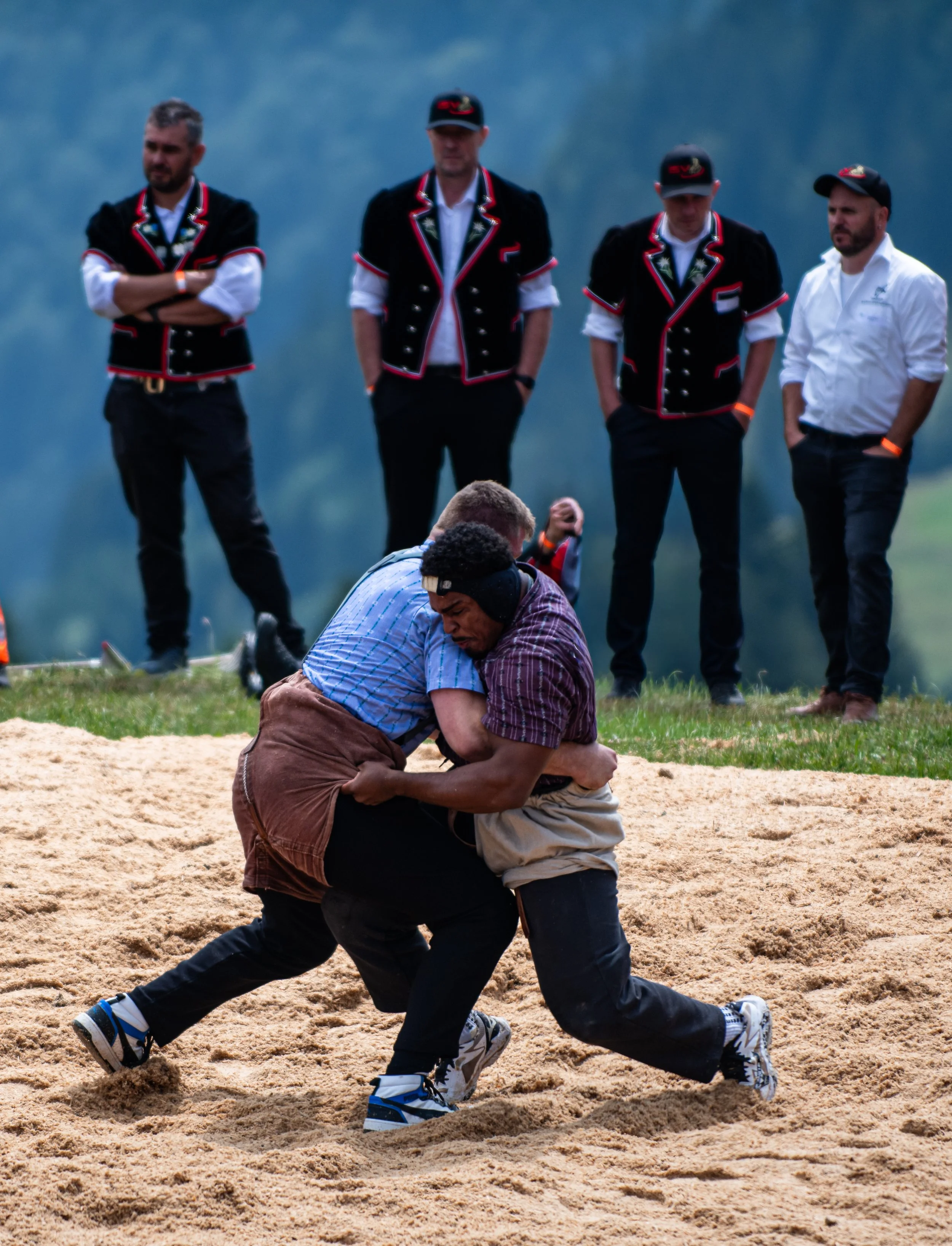 Zwei Männer in traditionellen Trachten, die bei einem Wrestling-Wettkampf auf einer Sandfläche miteinander kämpfen, während drei andere Männer im Hintergrund zuschauen.