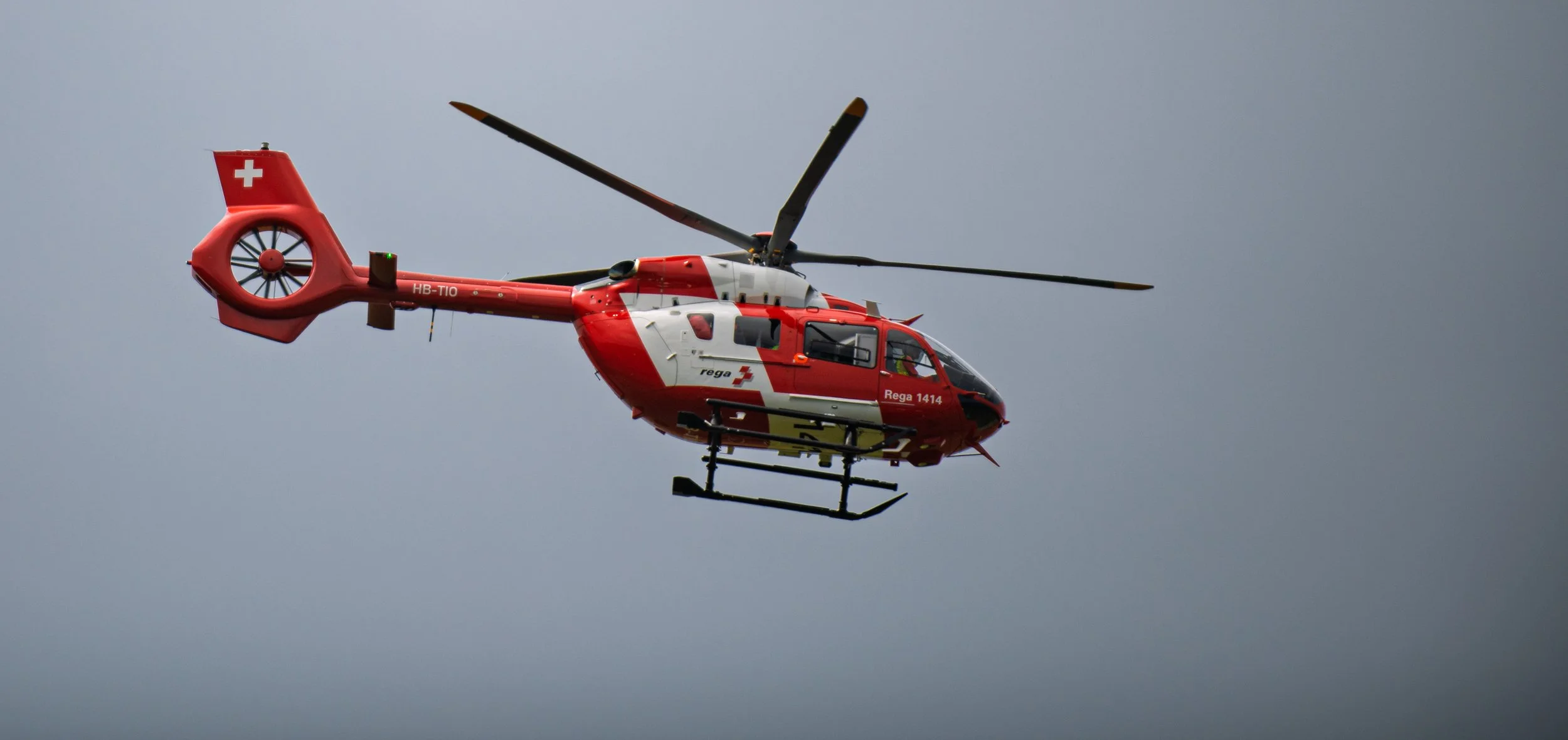 Rotes und weißes Rettungshubschrauber fliegt am Himmel, Wolken im Hintergrund.