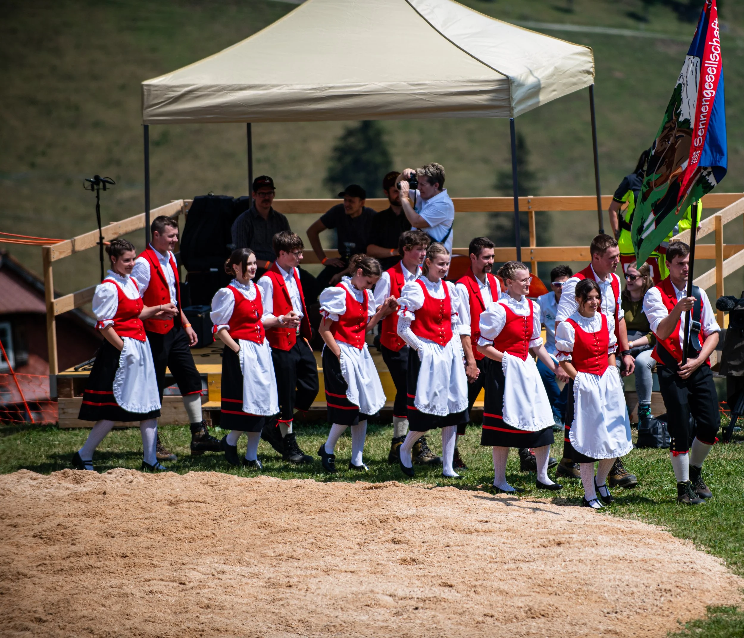 Gruppierung von Menschen in traditionellen Trachten, die bei einem Fest oder einer Veranstaltung im Freien stehen, zwei Frauen tragen rot-weiße Kleider, Männer in dunklen Hemden, im Hintergrund ein Dach und bunte Fahnen.