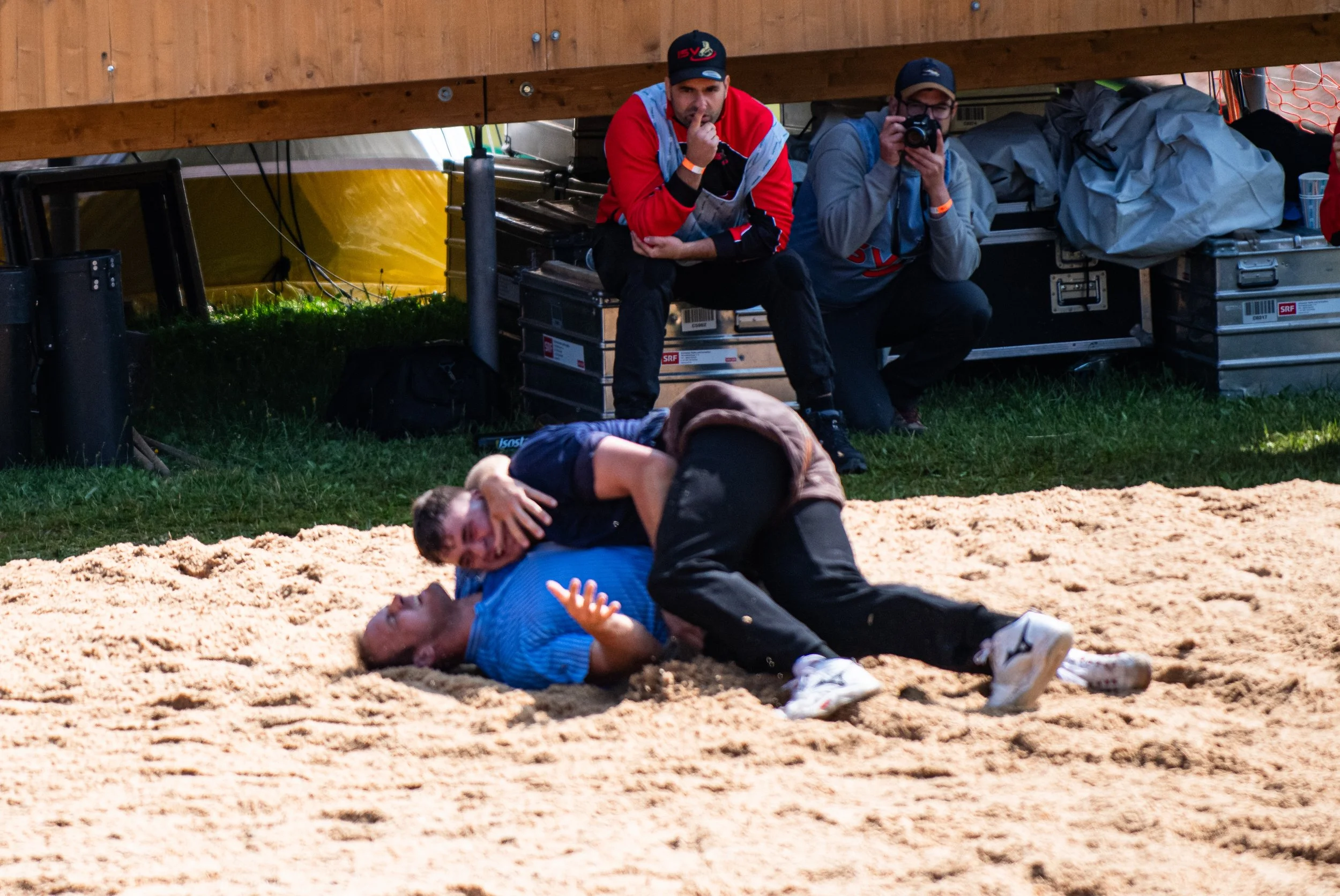 Zwei Menschen kämpfen in einem Wrestling- oder Ringenreitwettbewerb auf einem Sandboden, während zwei andere Personen im Hintergrund beobachten, eine davon filmt. Eine Person liegt auf dem Rücken, während die andere im Begriff ist, sie festzuhalten.