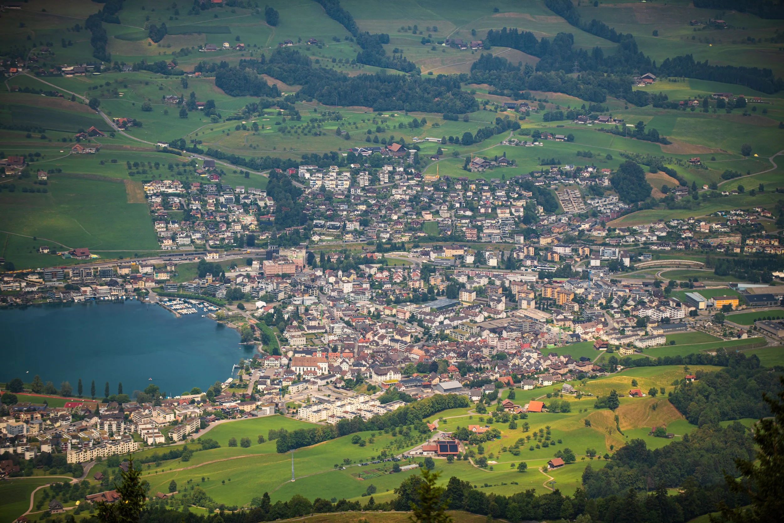 Luftaufnahme eines Hügellands mit Stadt und Wasser im Vordergrund, grüne Felder und Wälder im Hintergrund.