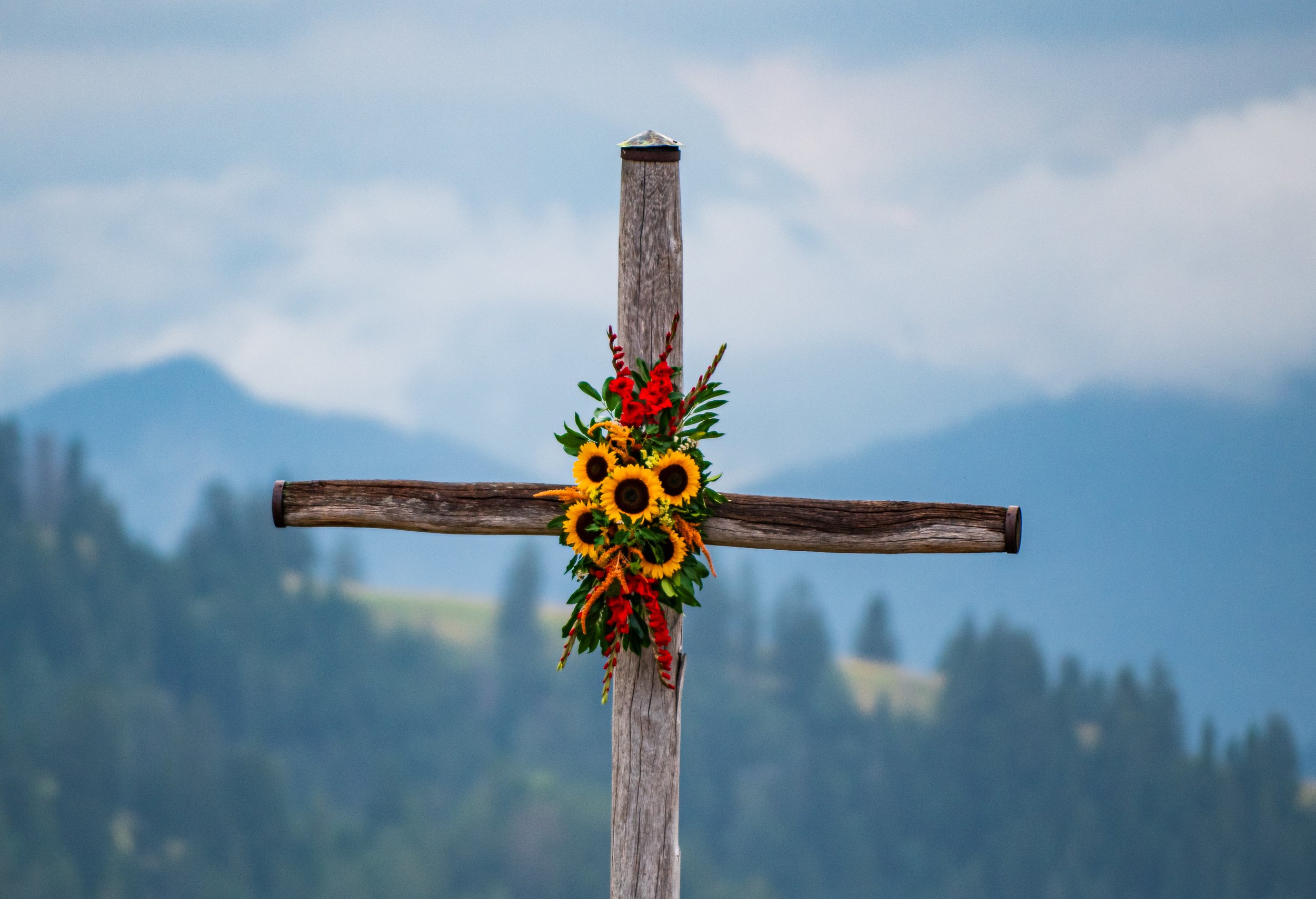 Holzkreuz mit bunten Blumen und Sonnenblumen vor nebligem Bergpanorama.