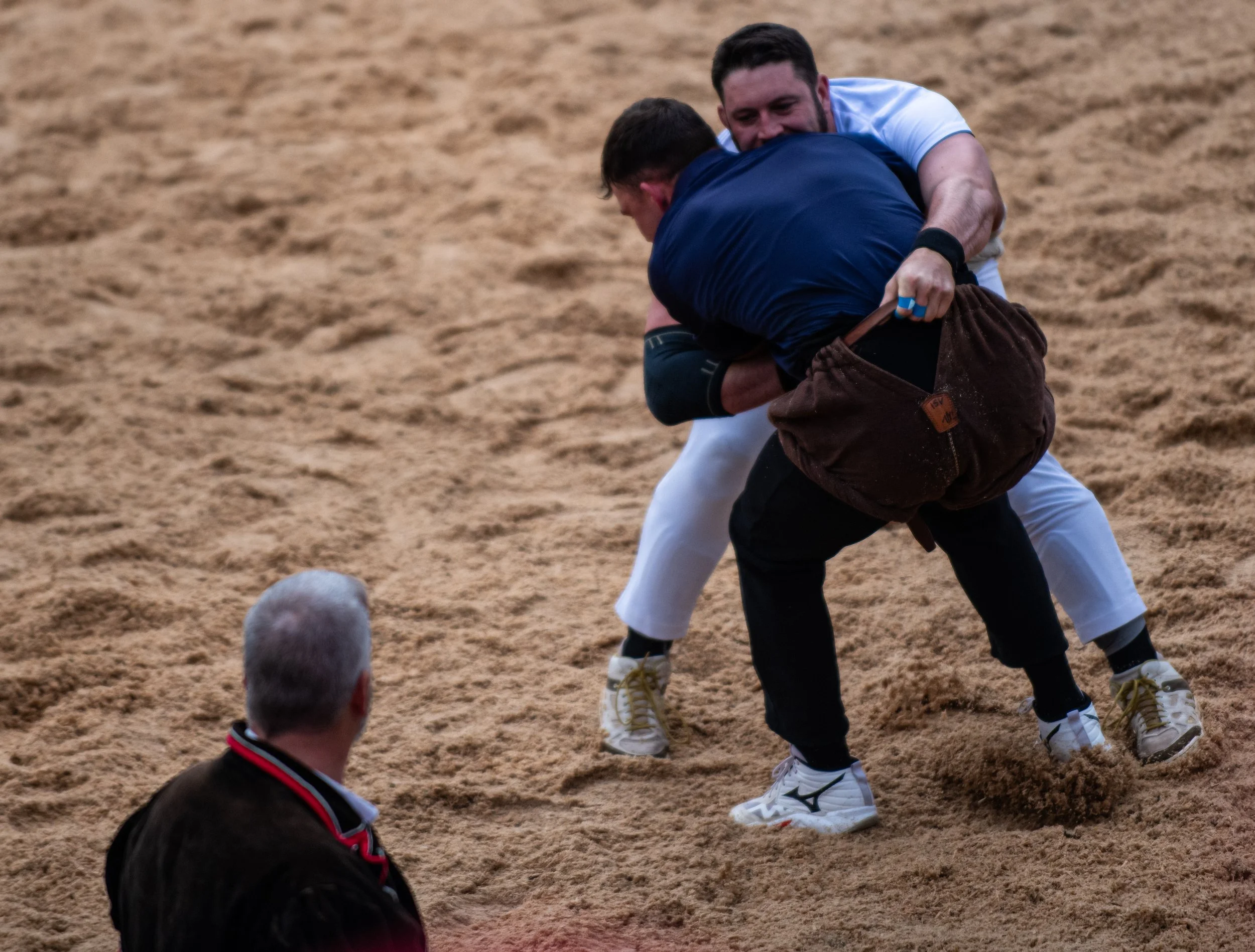 Zwei Männer im grappling-Stil konkurrenzieren auf einem Sandboden, einer in einem weißen T-Shirt und der andere in einem schwarzen Judo-Gi und braunen Gürtel.