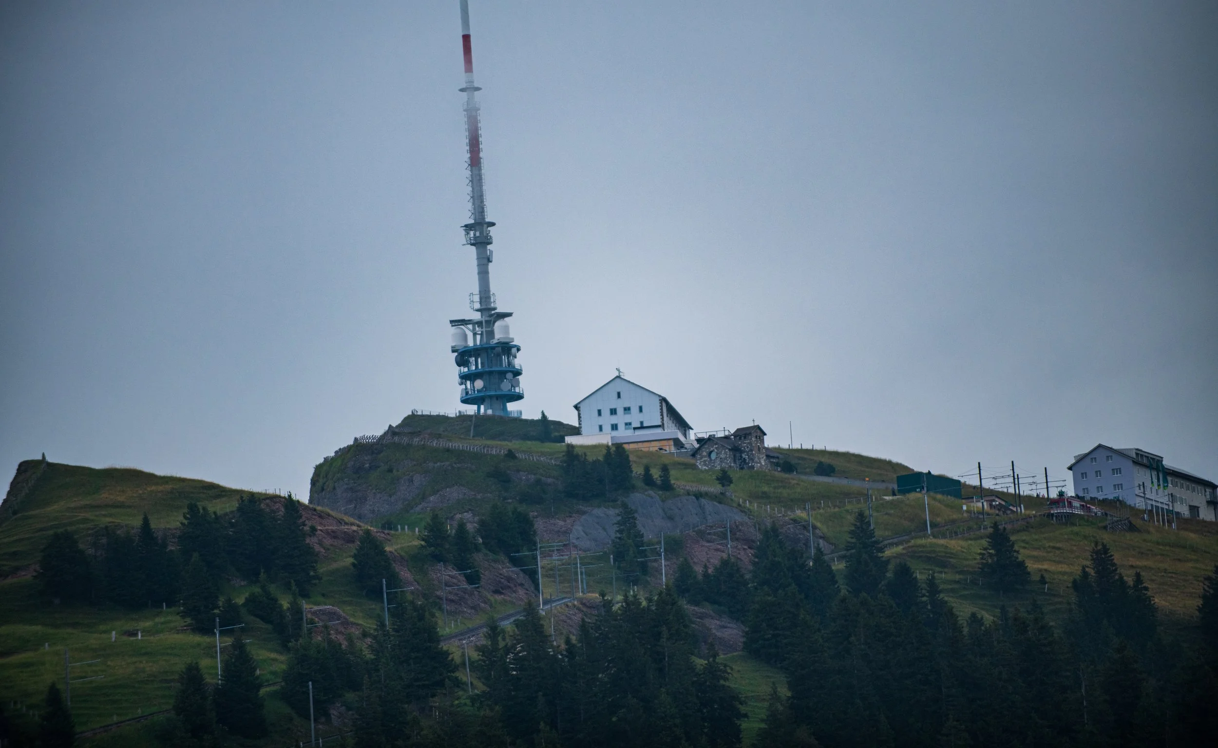 Ein Funkturm auf einem Hügel mit einigen Gebäuden im Vordergrund, umgeben von Bäumen und Wiesen, bei bewölktem Himmel.