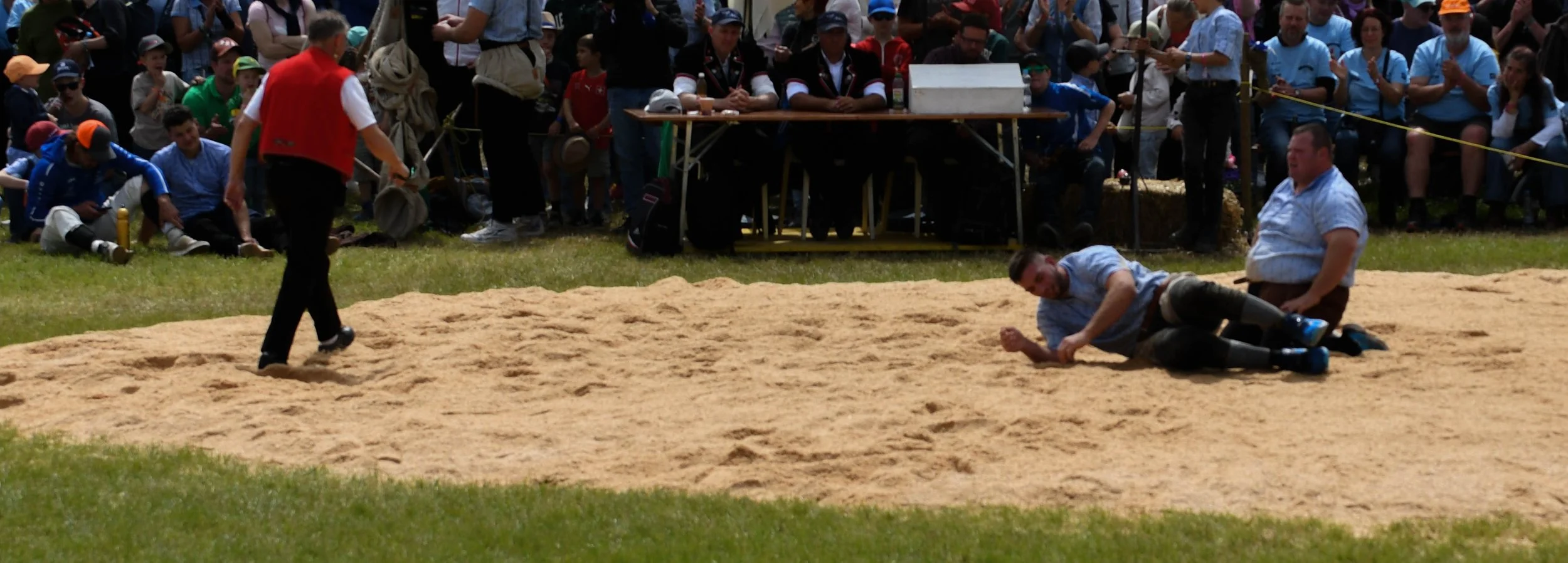 Zwei Männer beim Weitsprung, einer liegt auf dem Boden und der andere steht daneben, im Hintergrund eine Menschenmenge bei einem Sportevent.