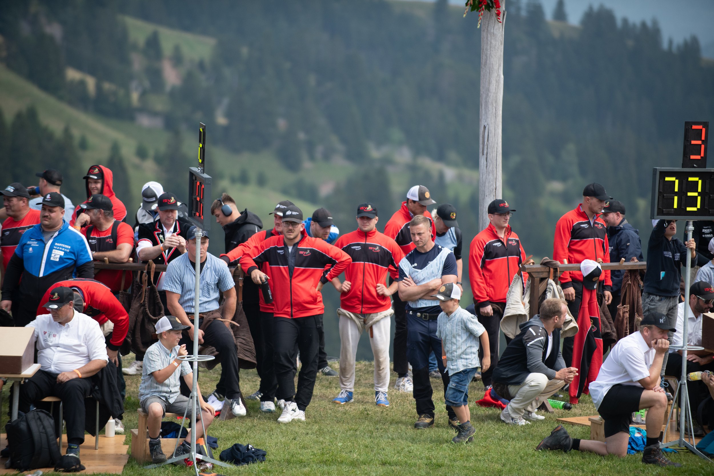 Viele Menschen, einige sitzend, andere stehend, bei einem Outdoor-Event auf einer Wiese mit Bergen im Hintergrund. Es gibt eine Anzeigetafel mit Zahlen. Die meisten tragen schwarze, rote oder bläulich-weiße Kleidung und Caps, einige hören Kopfhörer, 