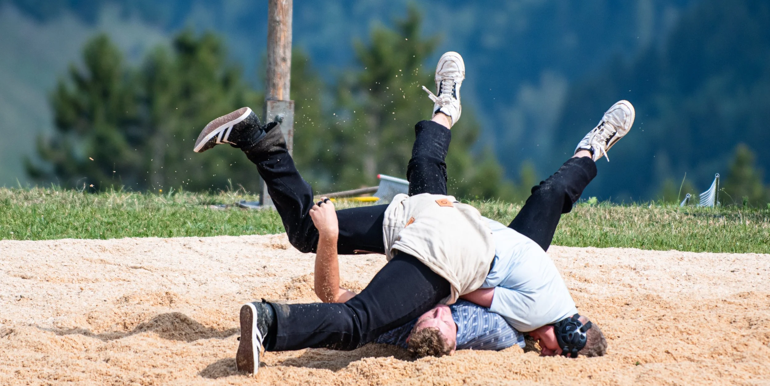 Zwei junge Männer fallen auf einer sandigen Feldbank während eines Spiels. Einer liegt auf dem Boden, der andere ist in der Luft, scheinbar inmitten eines Sturzes oder eines alpenangreifenden Spiels. Im Hintergrund sind Bäume und ein blauer Himmel si