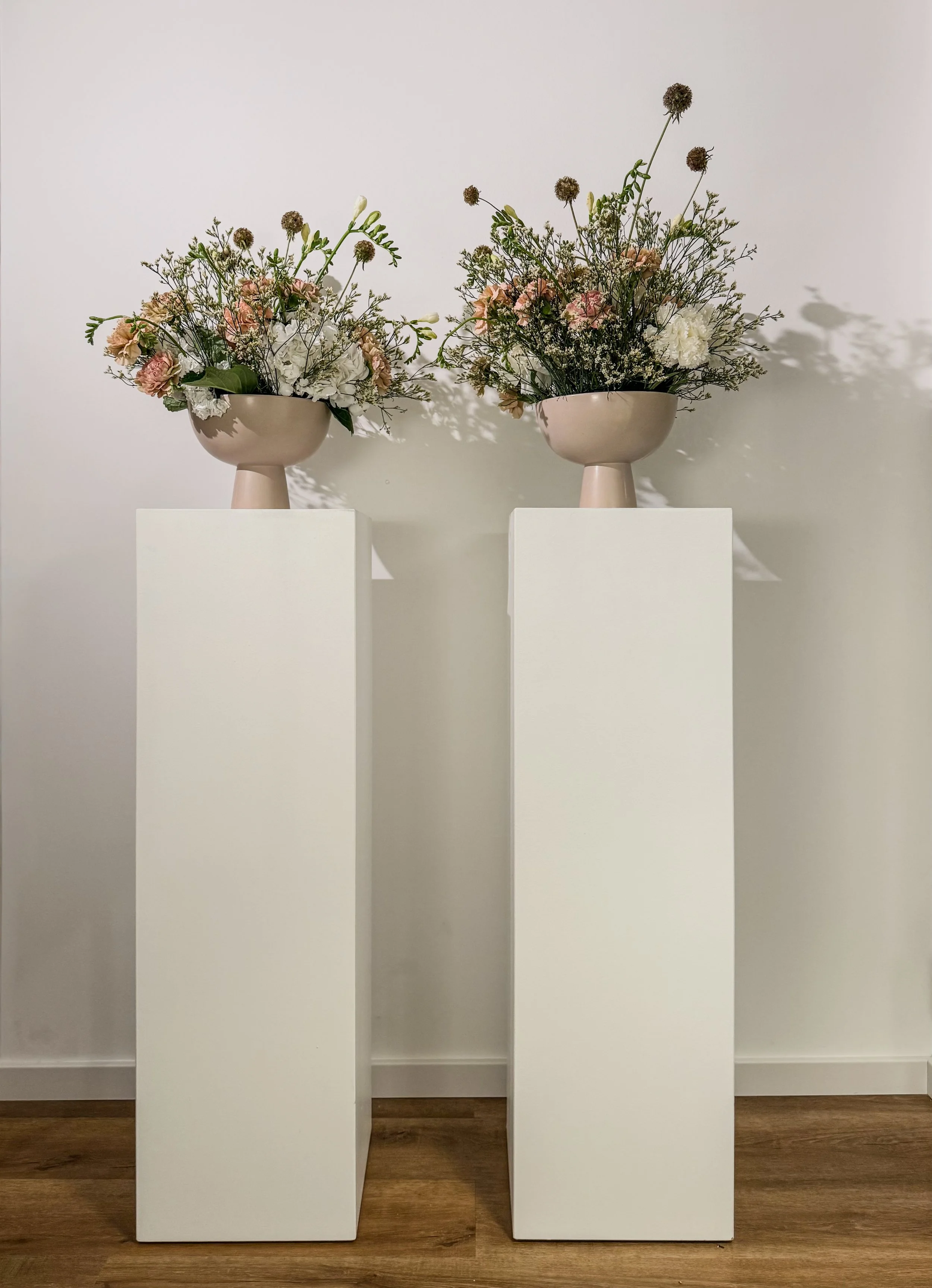 Zwei Blumenvasen mit Blumen auf weißen Podesten vor einer weißen Wand.
