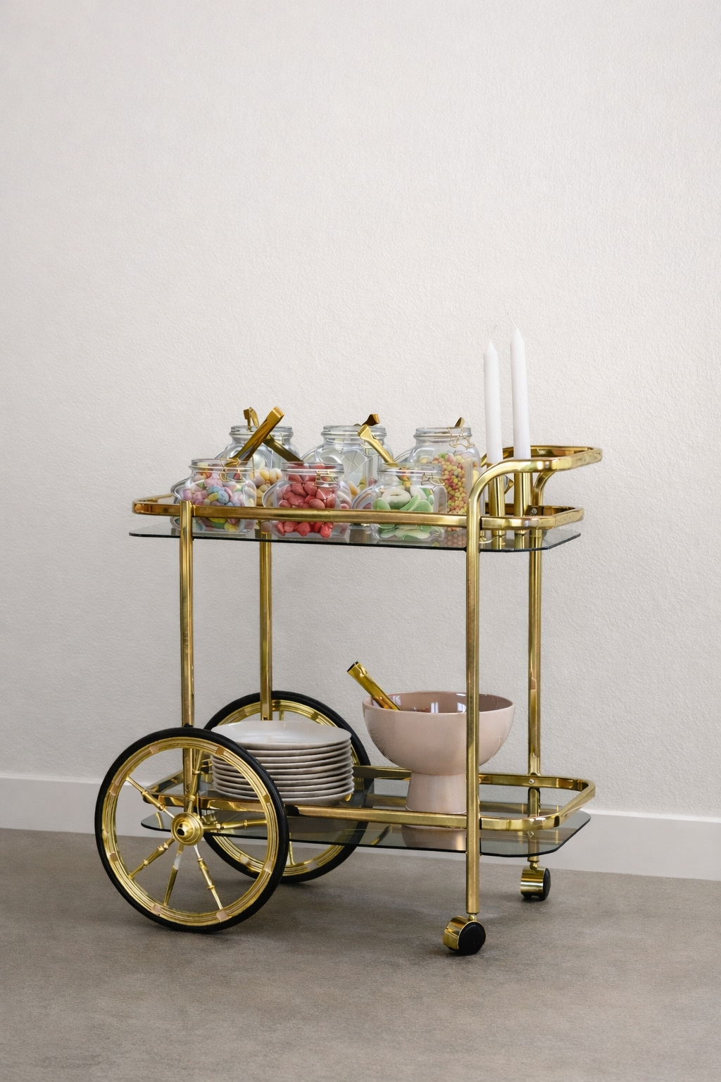 Goldfarbener Servierwagen mit Süßigkeiten in Gläsern, weißen Kerzen, Geschirr und Löffeln vor einer weißen Wand.