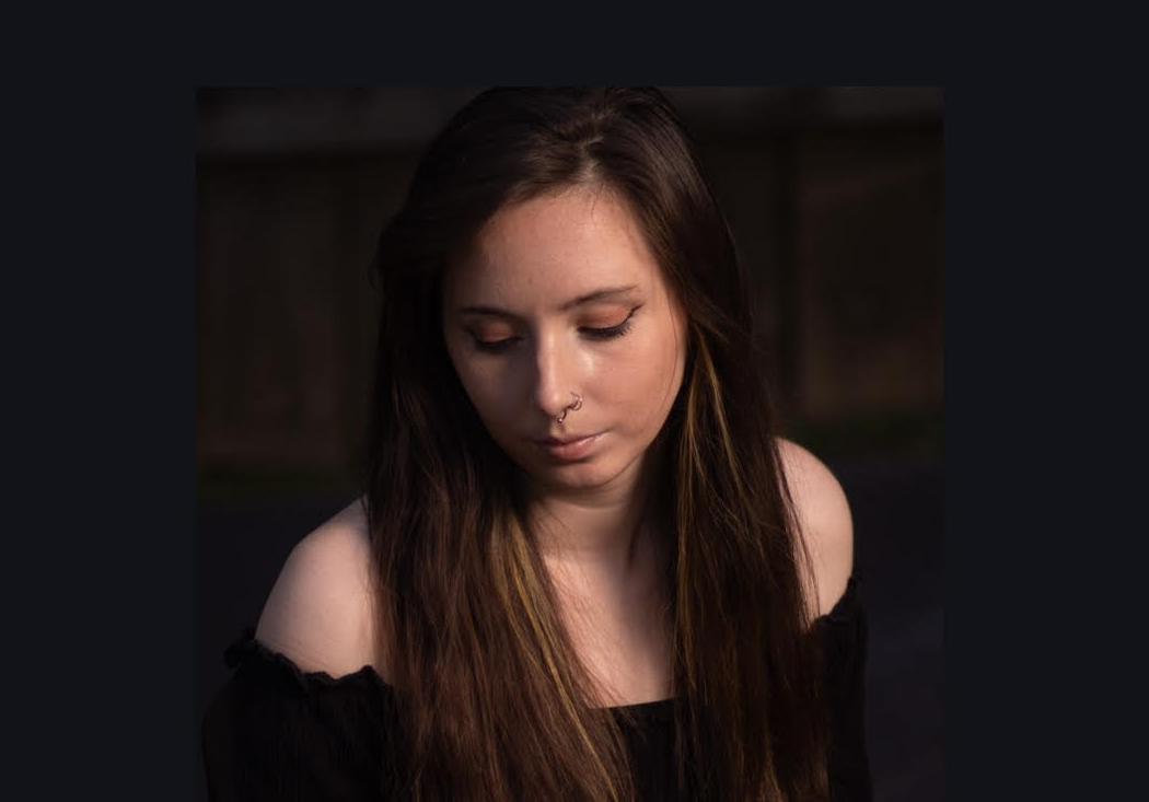 A young woman with long brown hair, wearing a black off-the-shoulder top, looking down with a contemplative expression.