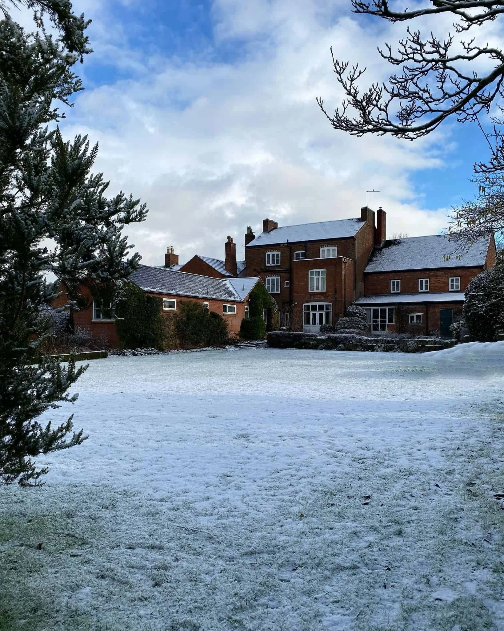 A snow-covered lawn in front of histroric listed red brick house with multiple chimneys, bay windows, crittall patio doors, detached garage, leafless trees, and a partly cloudy blue sky.