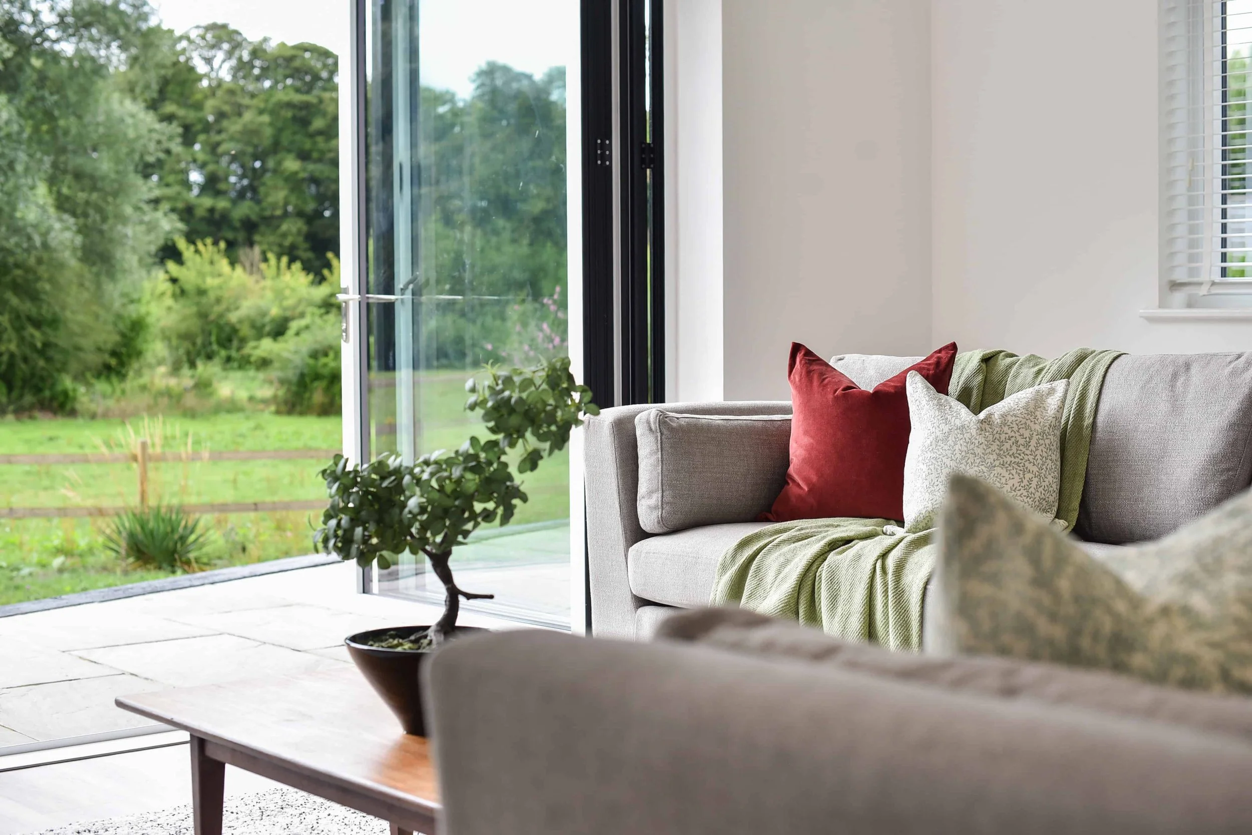 Living room with large bi-fold doors leading onto patio, vaulted ceiling, professional interior design with beige sofa, red and beige pillows, green throw blanket, potted plant