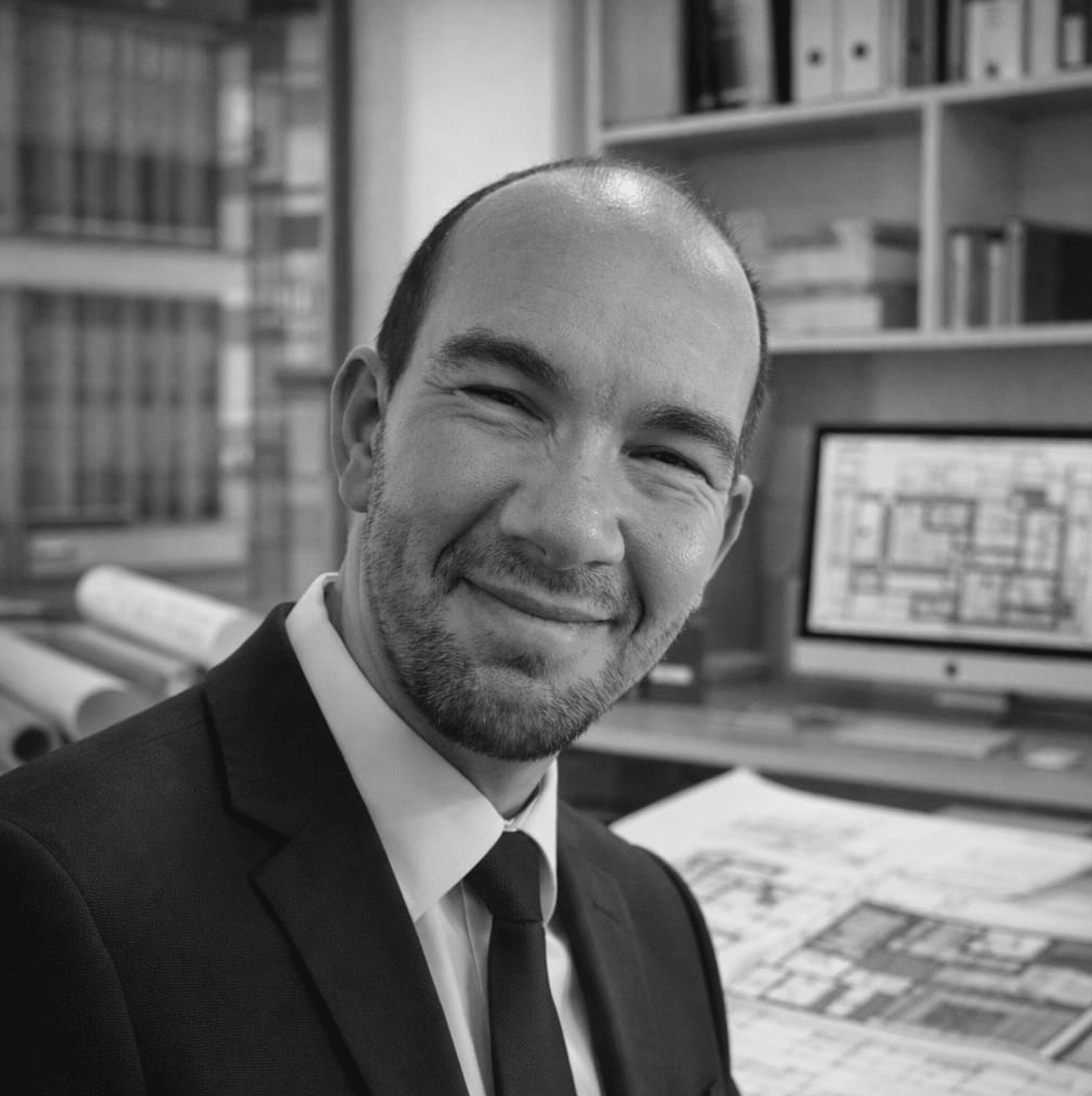 A man in a suit smiling in an office with architectural blueprints and a computer monitor displaying plans in the background, consultant at imj architecture.