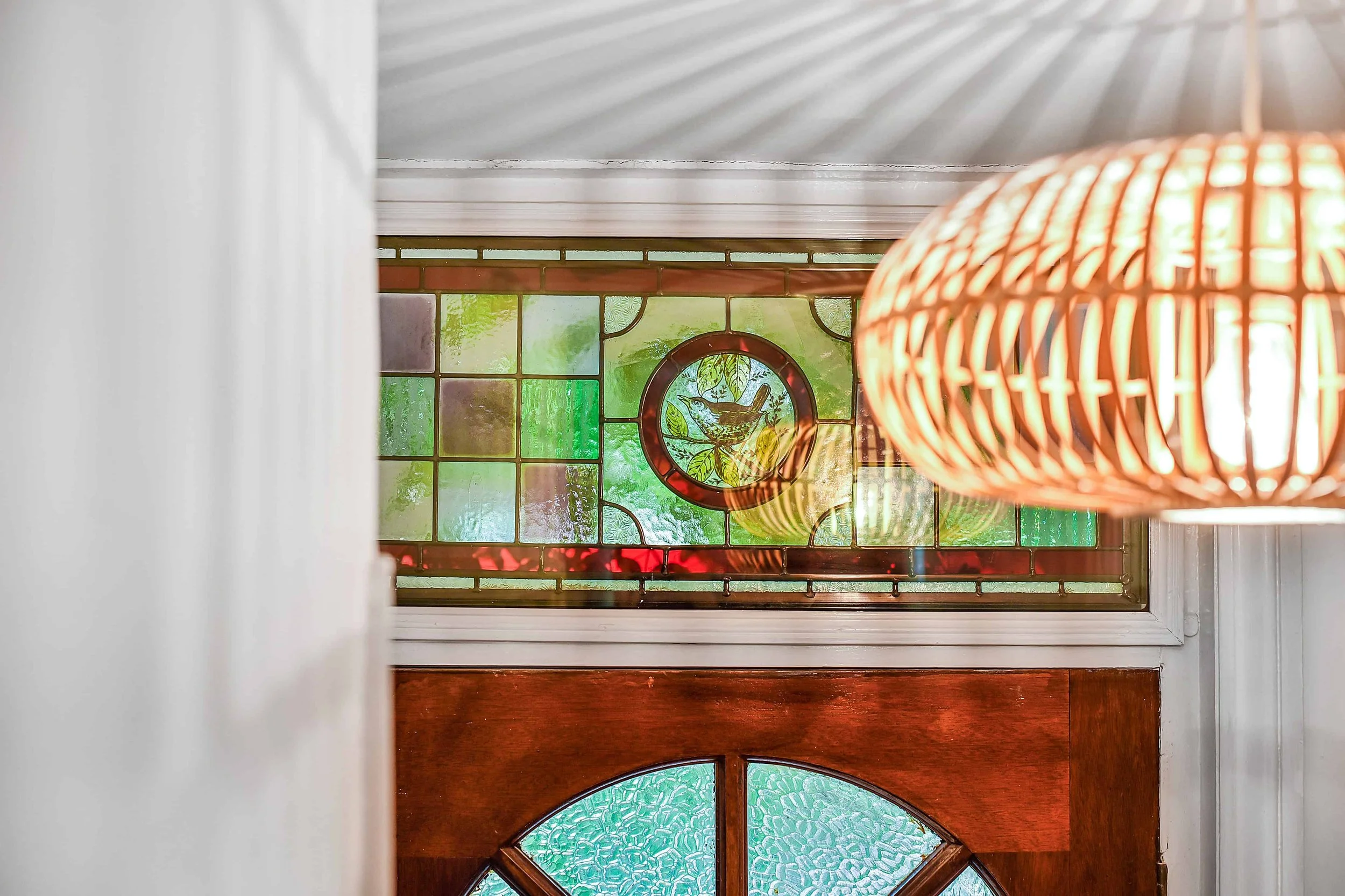 Traditional timber door with stained glass window, with a hanging wicker lampshade in the foreground, and white walls and trim surrounding the window.
