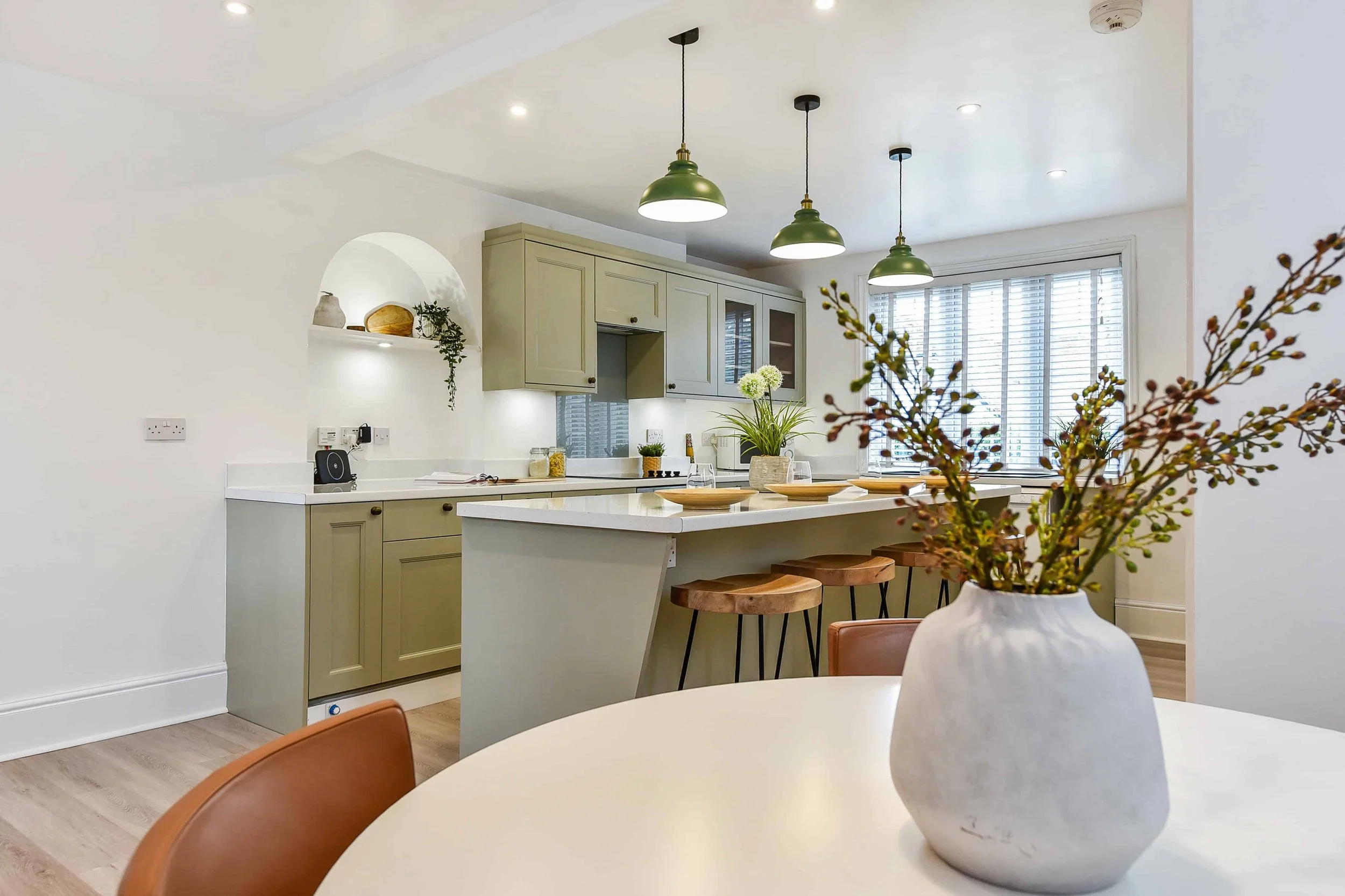 Traditional shaker style kitchen with hand painted pastel coloured cabinets, open plan dining room with table, granite kitchen worktops, Belfast sink and a window with blinds.. Green pendant lights hang over the island.
