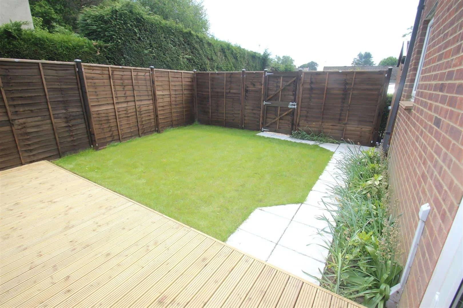 A refurbished rear garden in a rental property with a small grassy lawn, a wooden deck, a brick house on the right, and a brown wooden fence surrounding the yard.