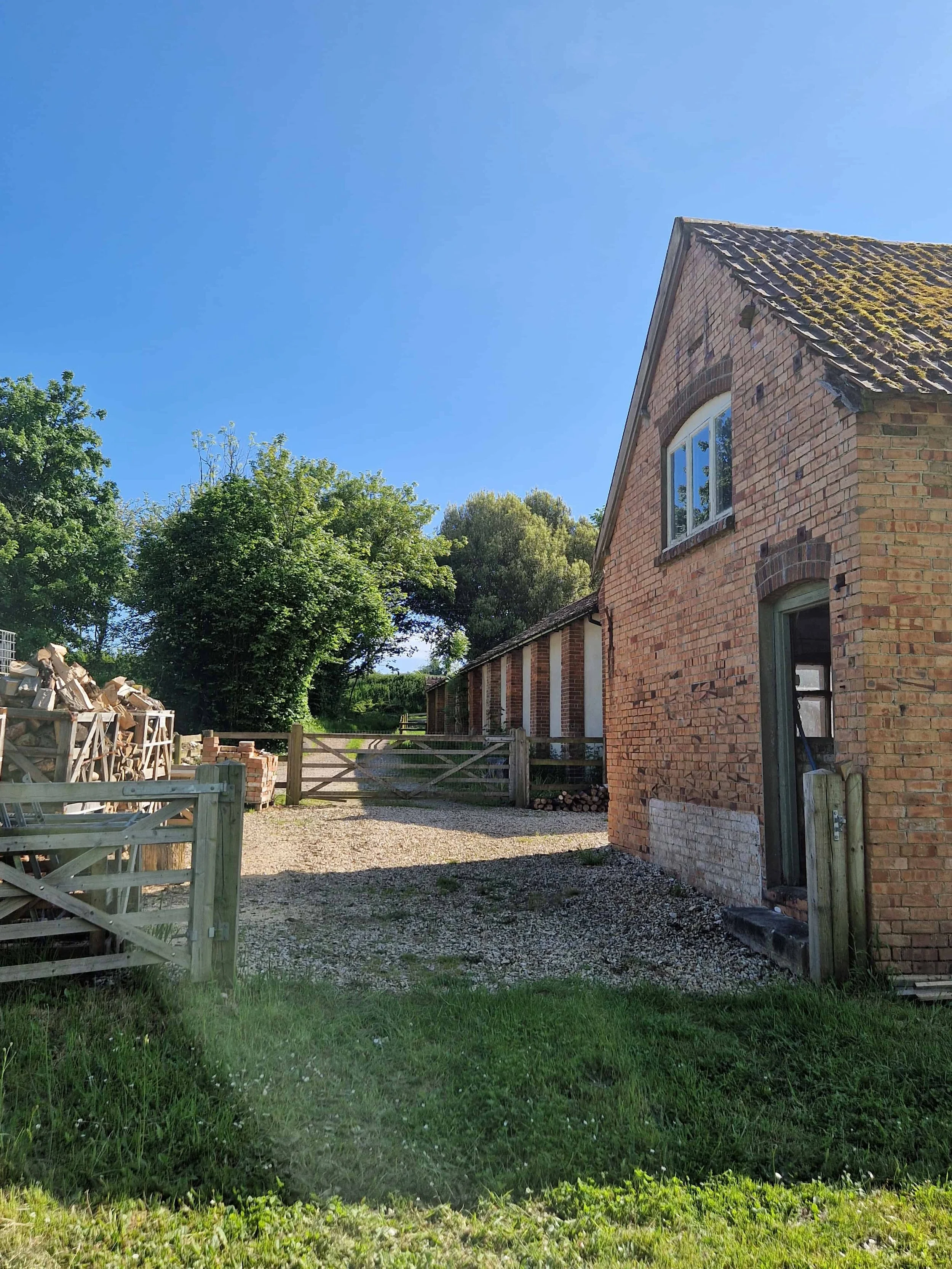 Brick barn in a conservation area with an arched window heads, tiled roof, surrounded by green trees, a wooden fence, and a pile of firewood on the left, under a clear blue sky.