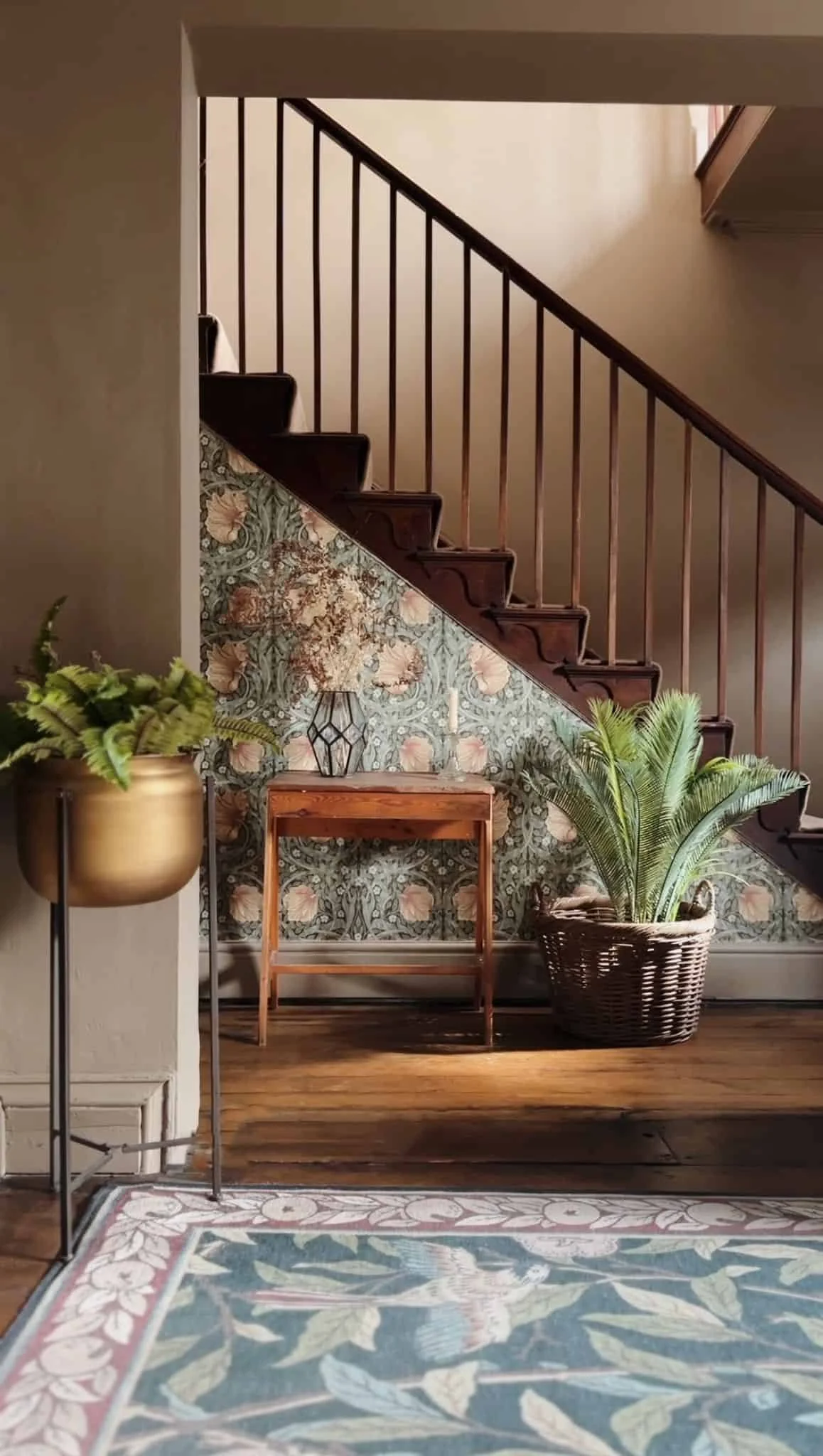 Original timber Georgian staircase with wooden steps and swept handrail and spindles, decorated with floral wallpaper in the background. In the foreground, there are plants in a gold pot and a wicker basket.