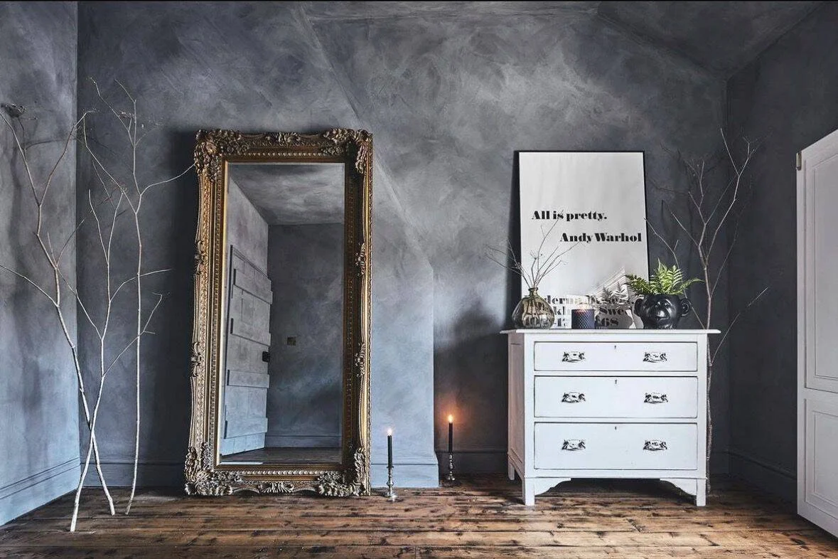 Interior of a room with a large ornate mirror, a white chest of drawers, framed art, candles, vases, and branches, with reclaimed wooden flooring and textured chalk paint grey walls.