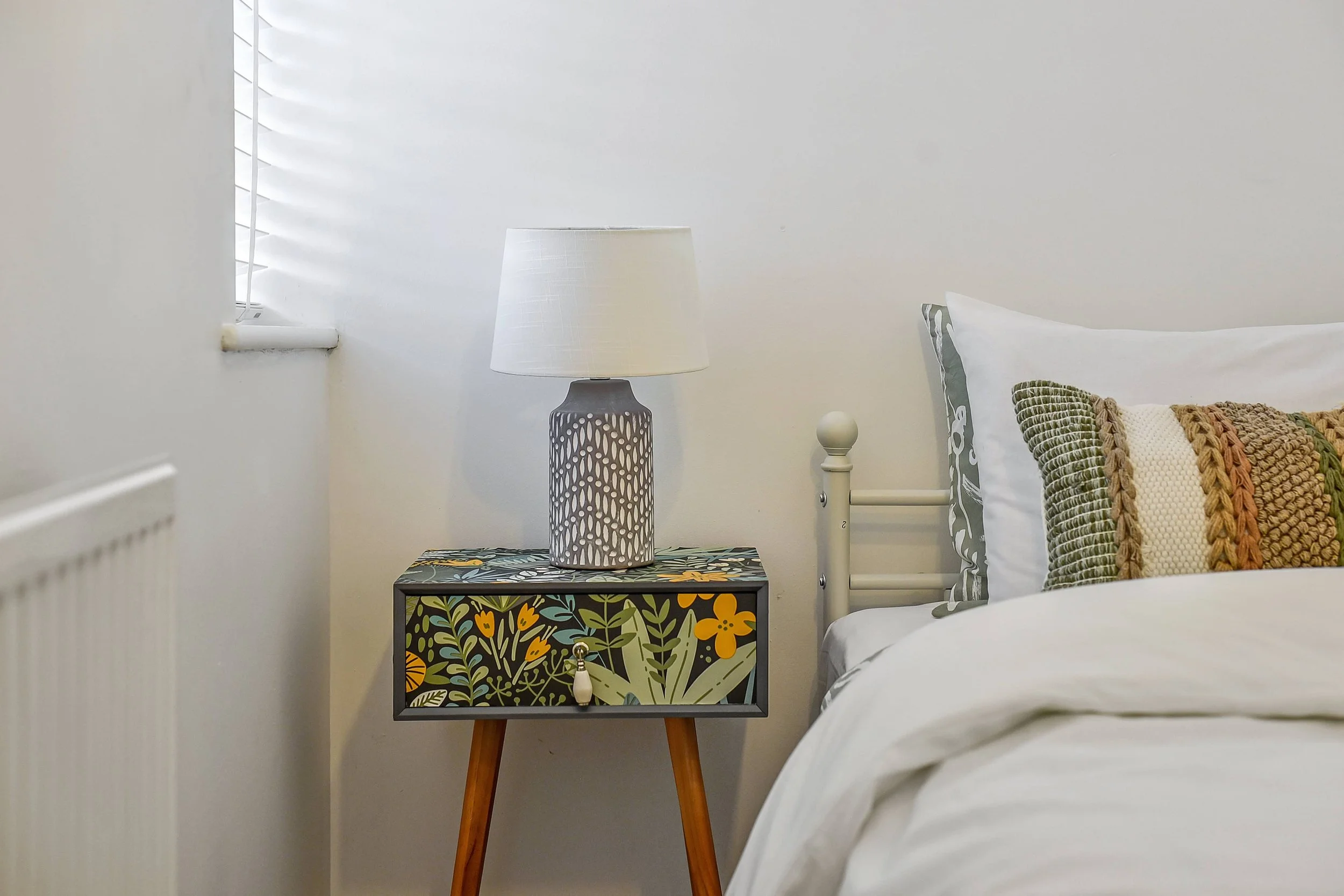 Bedroom interior design, corner with a bedside table, a patterned lampshade, a bed with chopped pillows, and blinds on a window, neutral colour scheme.